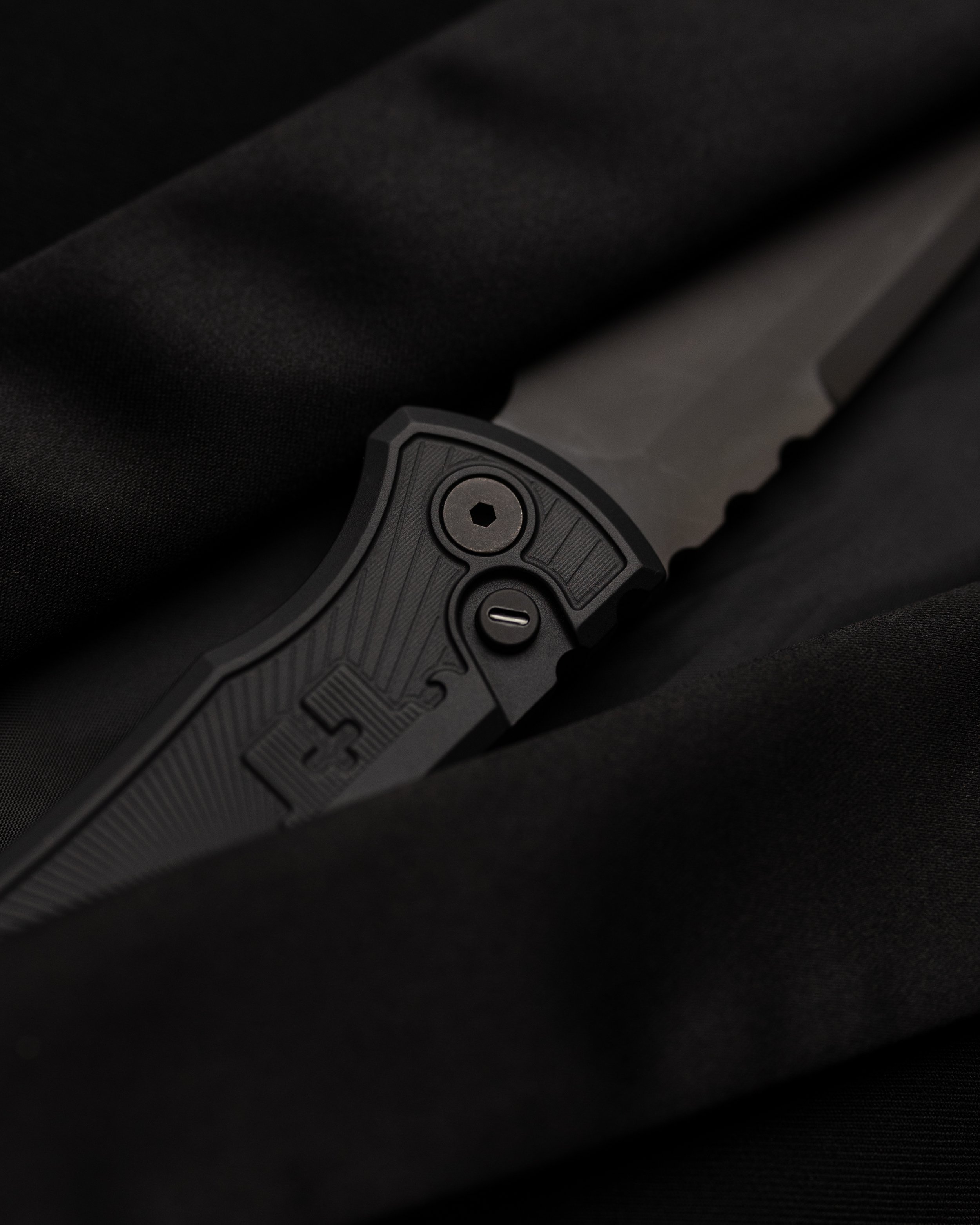 Close-up image of a tactical knife with a textured black grip and a partially visible blade, placed on black fabric.