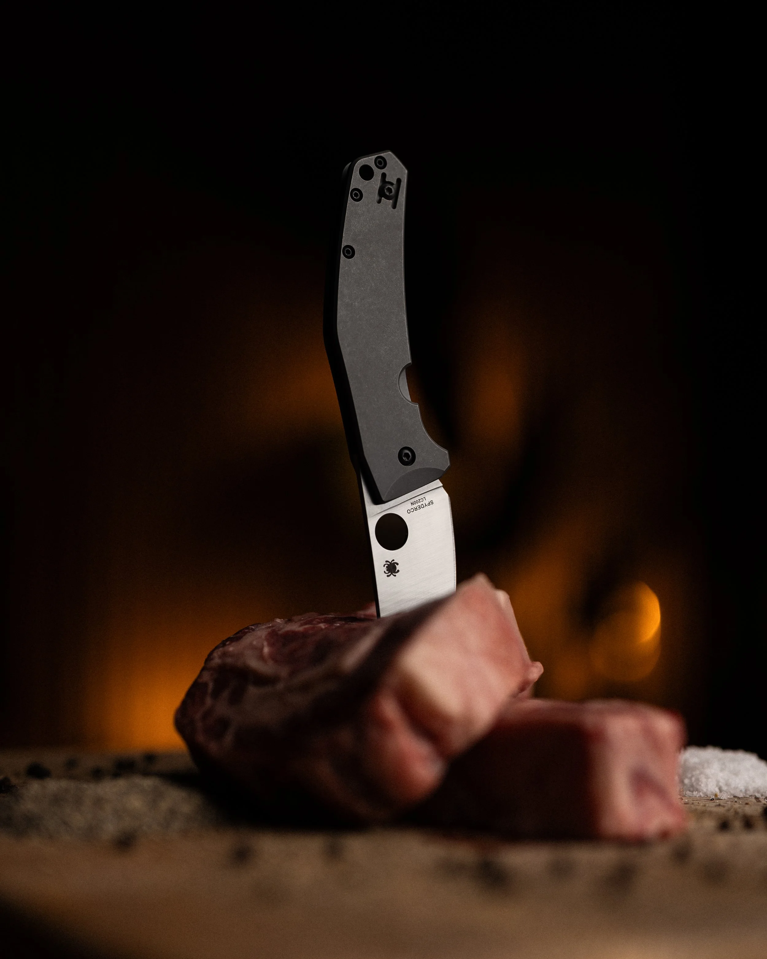 Close-up of a hand slicing a piece of raw meat with a Spyderco kitchen knife, against a dark background