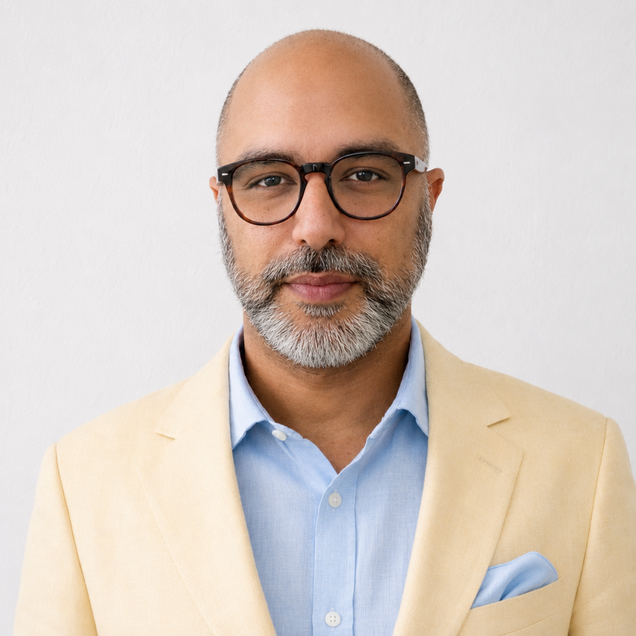 A middle-aged man with glasses, a beard, and a bald head posing against a plain white background. He is wearing a light yellow blazer over a light blue shirt with a pocket square.