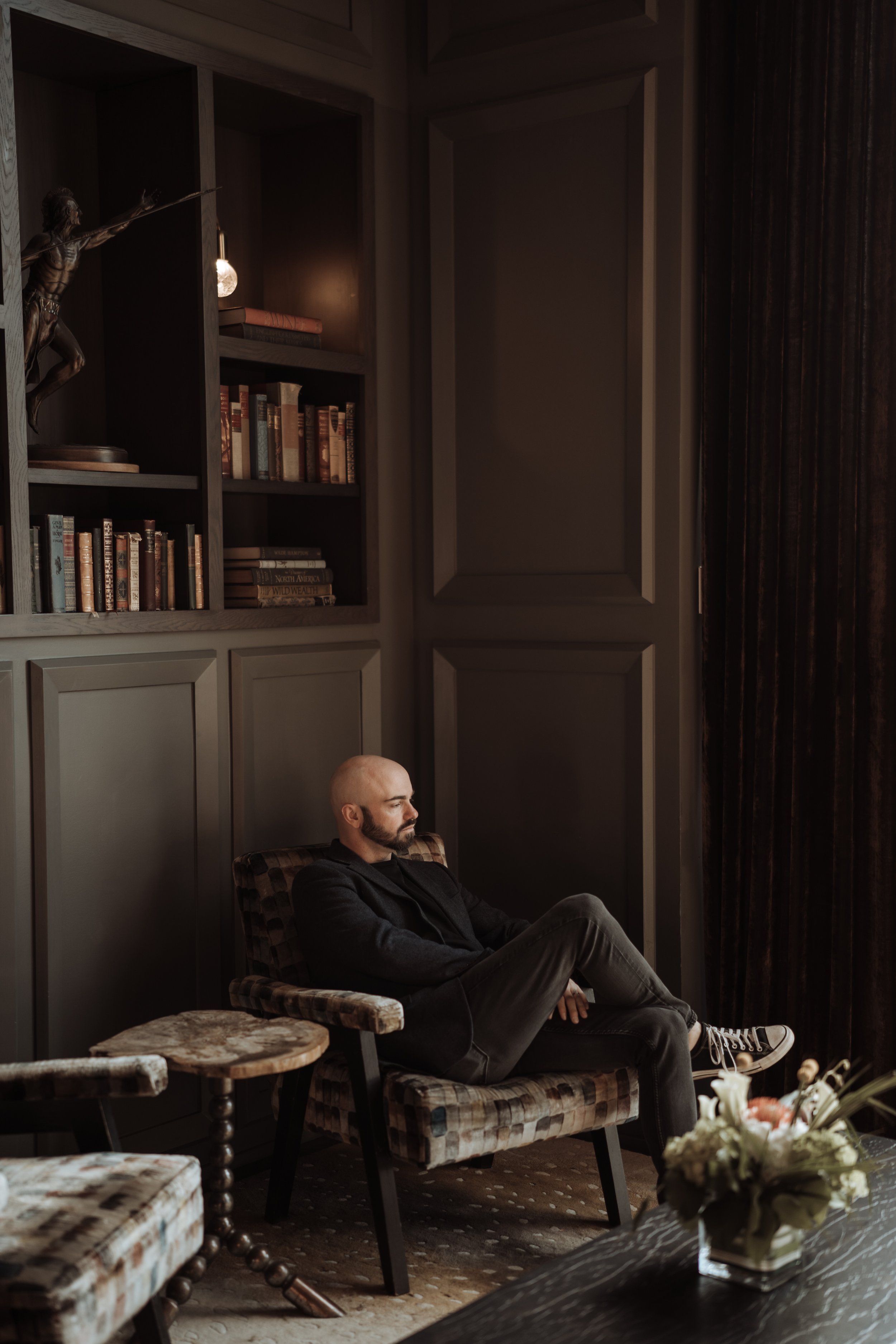A man with a shaved head and beard sitting in a patterned armchair in a dimly lit room, with bookshelves and dark curtains behind him, and a flower arrangement on a table in the foreground.