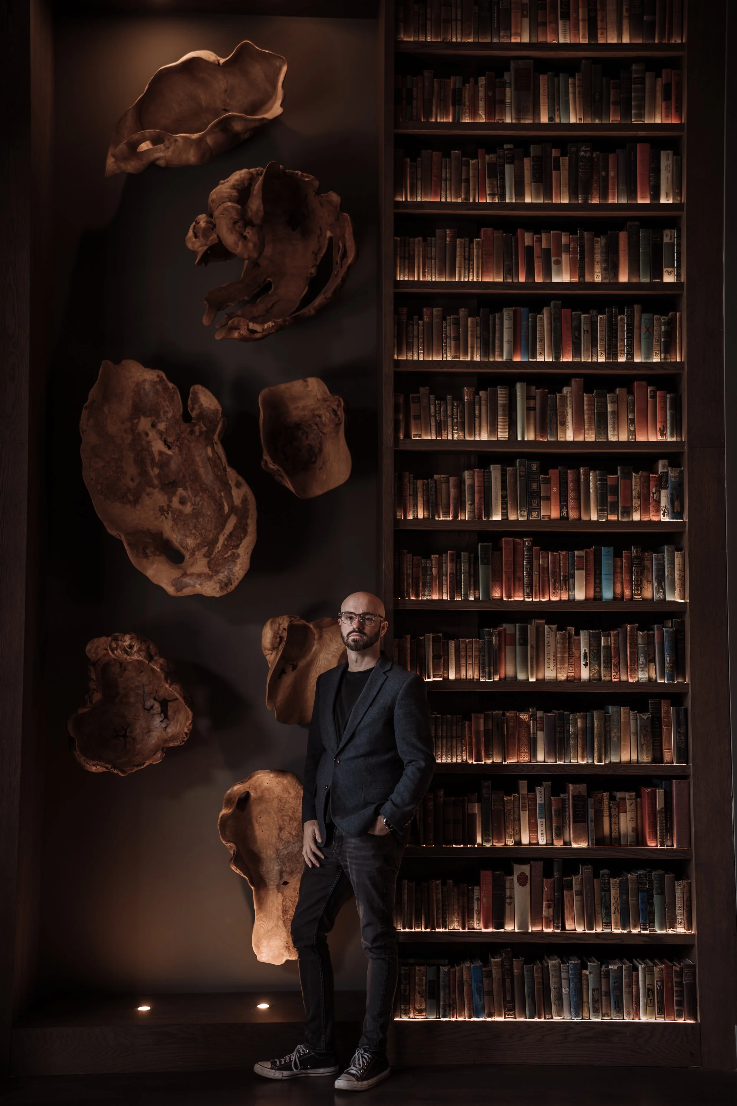 A man in a dark blazer and glasses leaning against a dark wooden bookshelf filled with books. The bookshelf is decorated with large, shaped wooden sculptures on the left side. The lighting is warm and ambient, creating a cozy atmosphere.