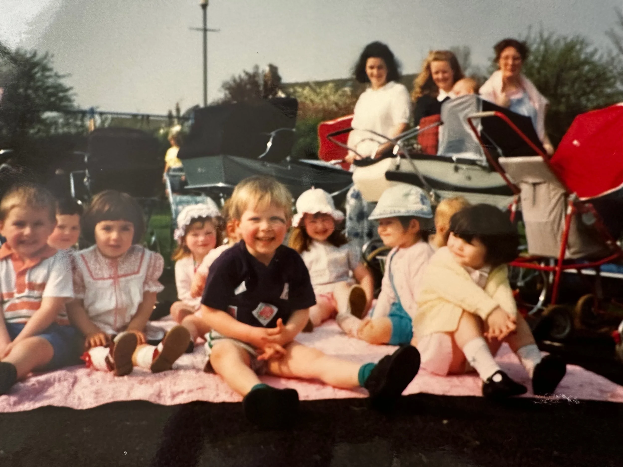 Children sitting on a pink blanket, smiling, with three women standing behind them near a vintage car during daytime.