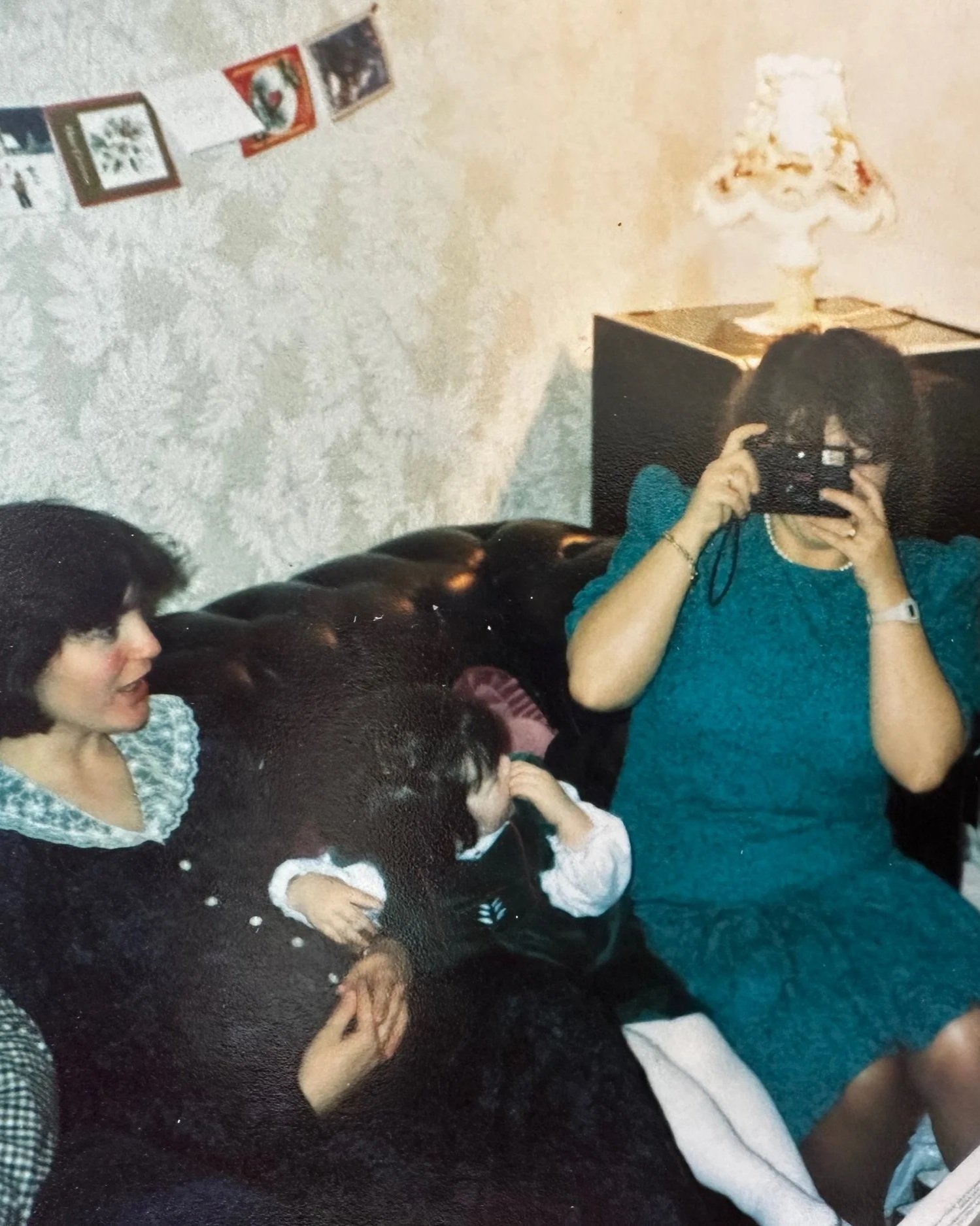 A woman with dark hair and glasses taking a mirror selfie in a living room with a lamp on a black table and a string of small pictures hanging on the wall, while two children sit on a black couch nearby.