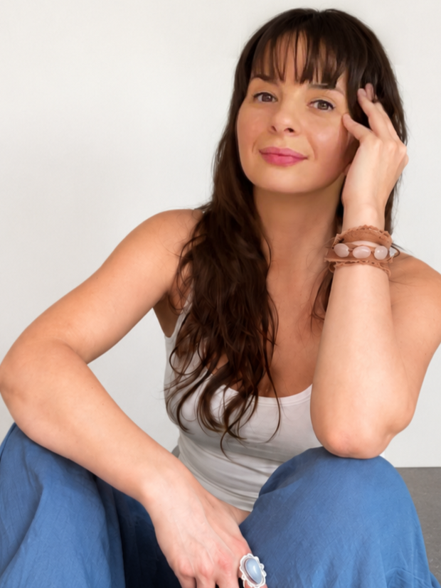 A woman with long brown hair and bangs, wearing a sleeveless white top and blue pants, sitting against a plain white background, smiling gently with her hand resting on her face.