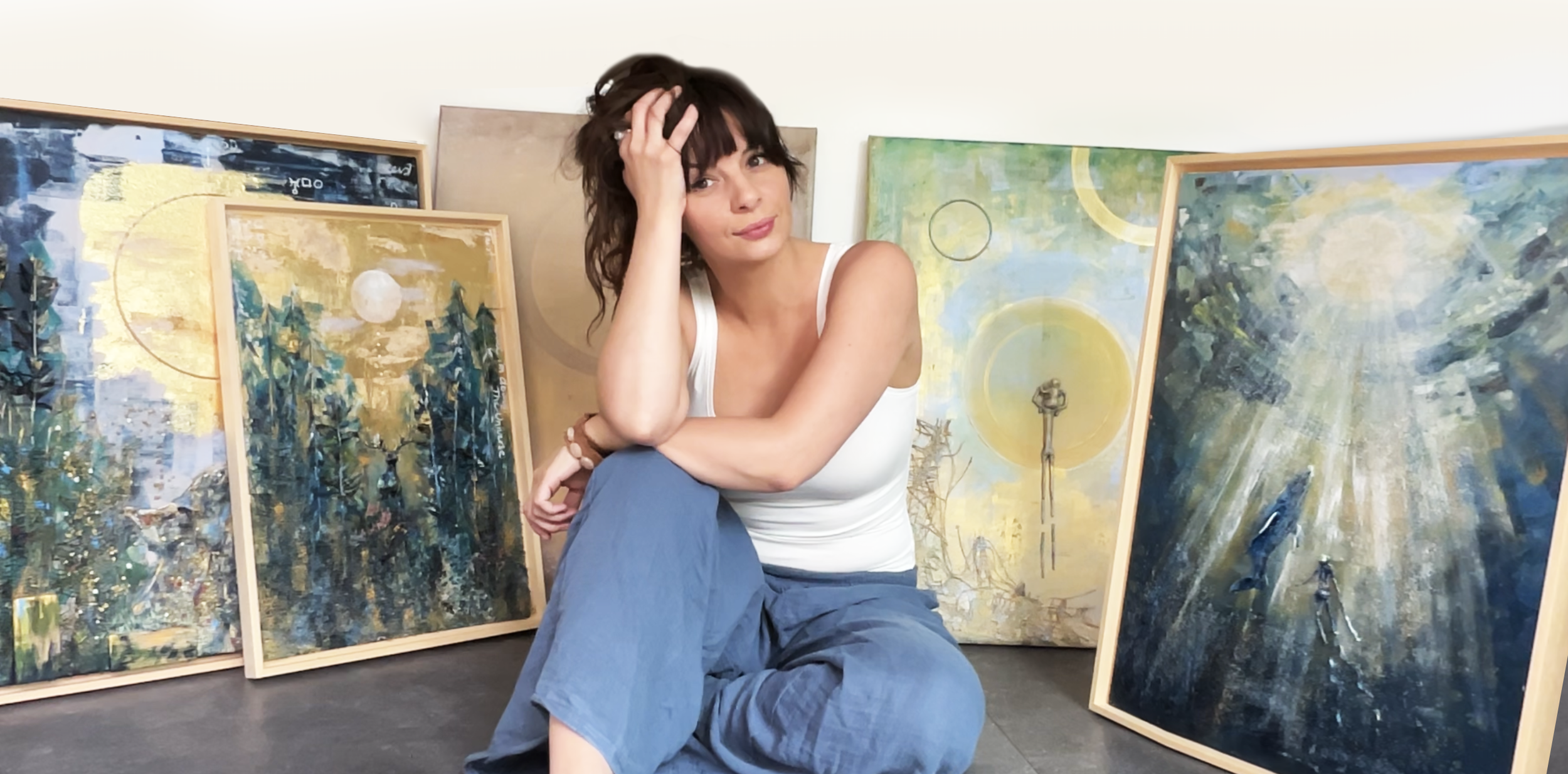 A woman sitting on the floor in front of several abstract paintings on display, with one hand on her head and a relaxed expression.