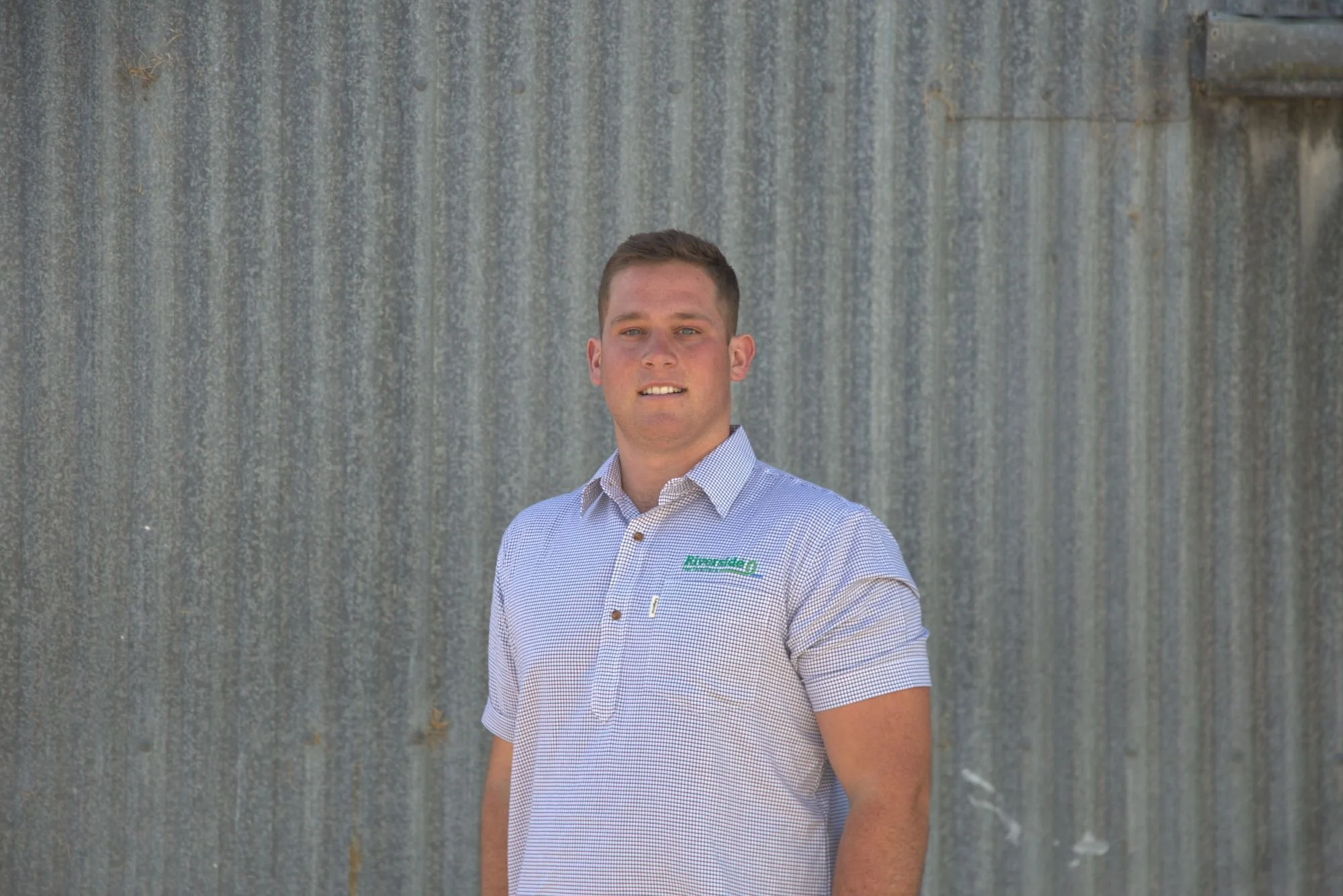 A young man with short brown hair wearing a light blue and white checkered shirt with a logo that says 'Riverside' on the chest, standing against a weathered metal wall.