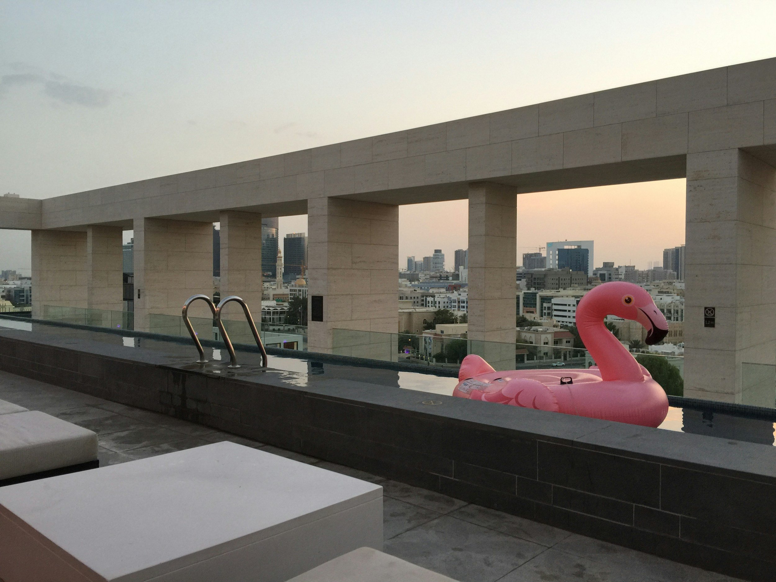 Luxury rooftop swimming pool with city skyline view, inflatable pink flamingo float, modern white seating, and a beige architectural structure with columns at sunset.