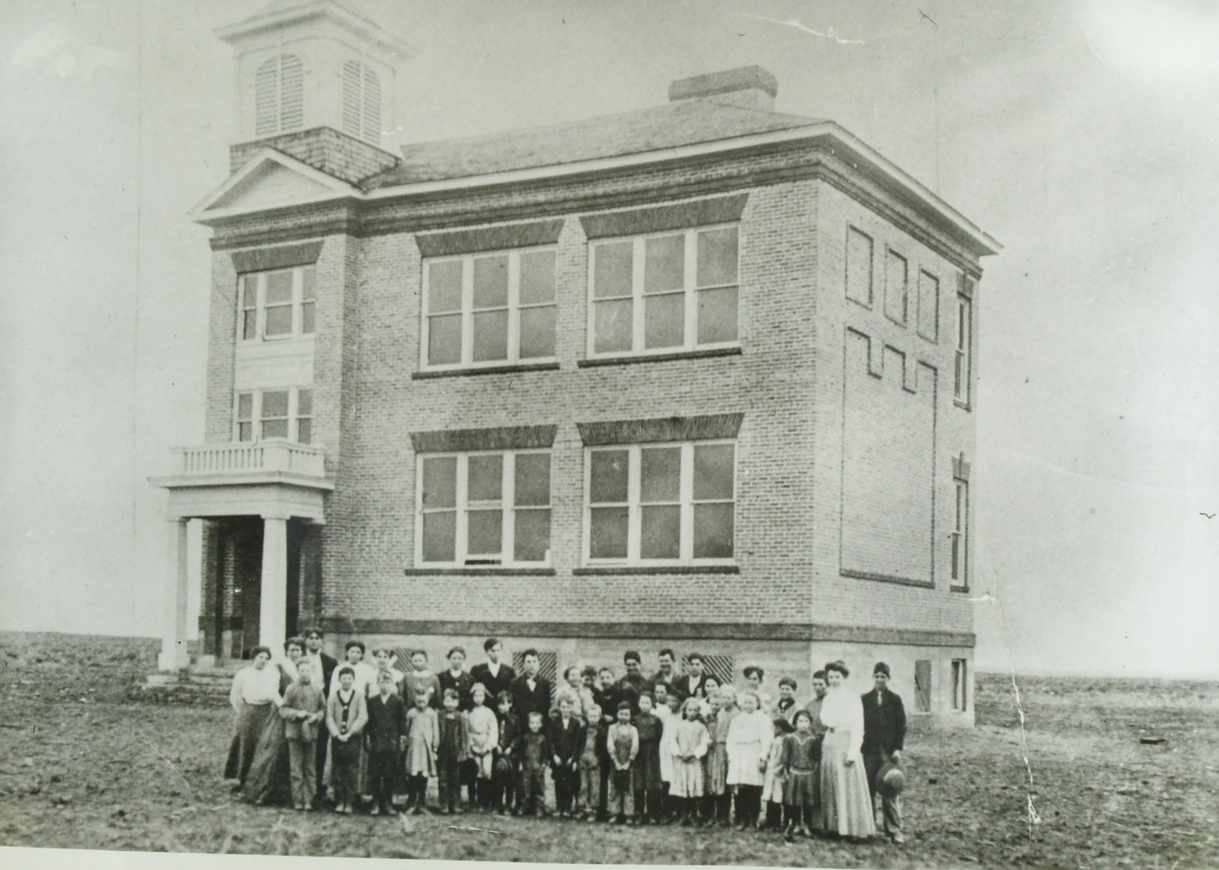 Black and white photo of a group of children and a few adults standing in front of a large brick building with multiple windows and a porch, possibly from the early 20th century.