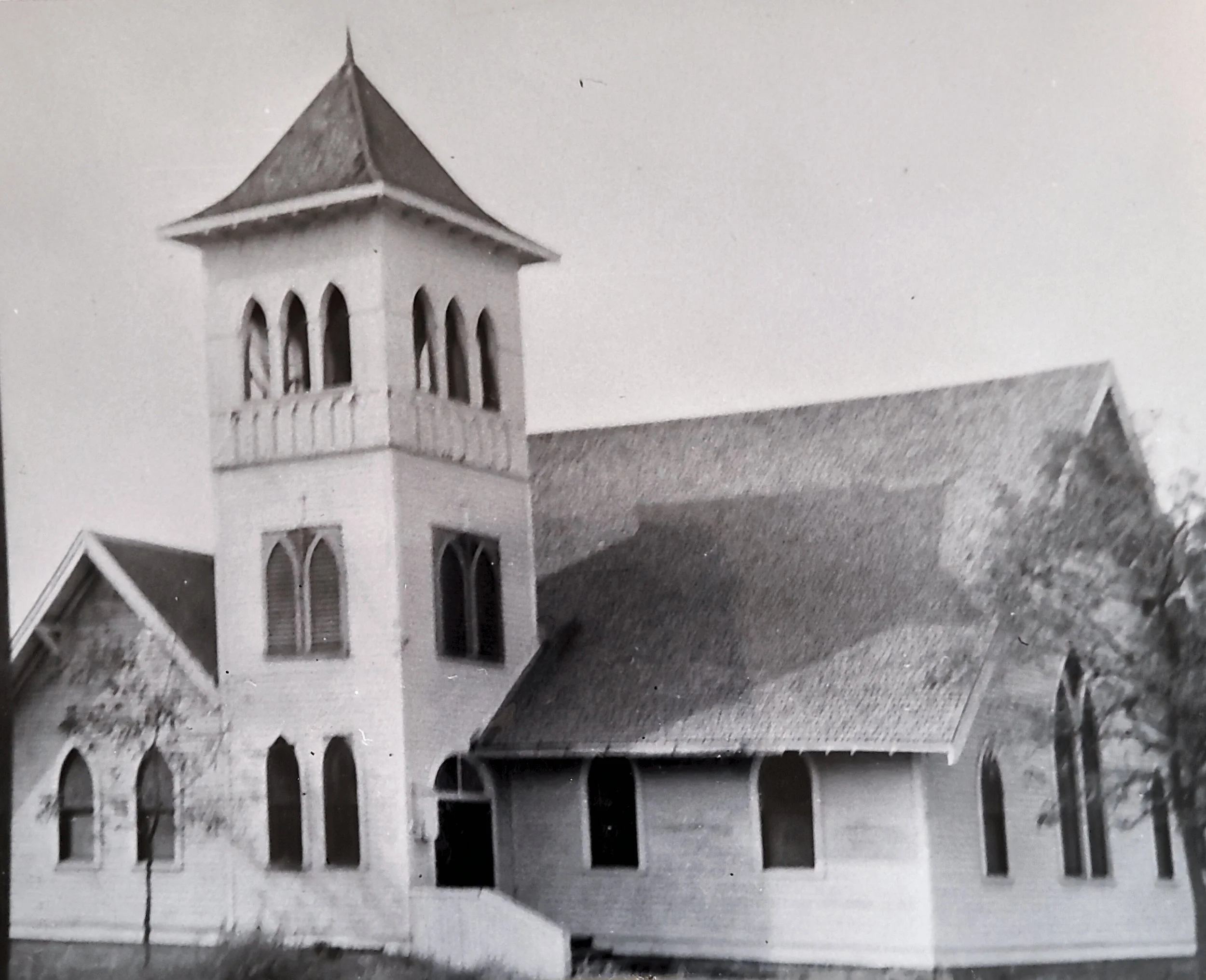 Black-and-white photo of a church tower with three arched windows and a pointed roof, with part of the church roof visible in the foreground.