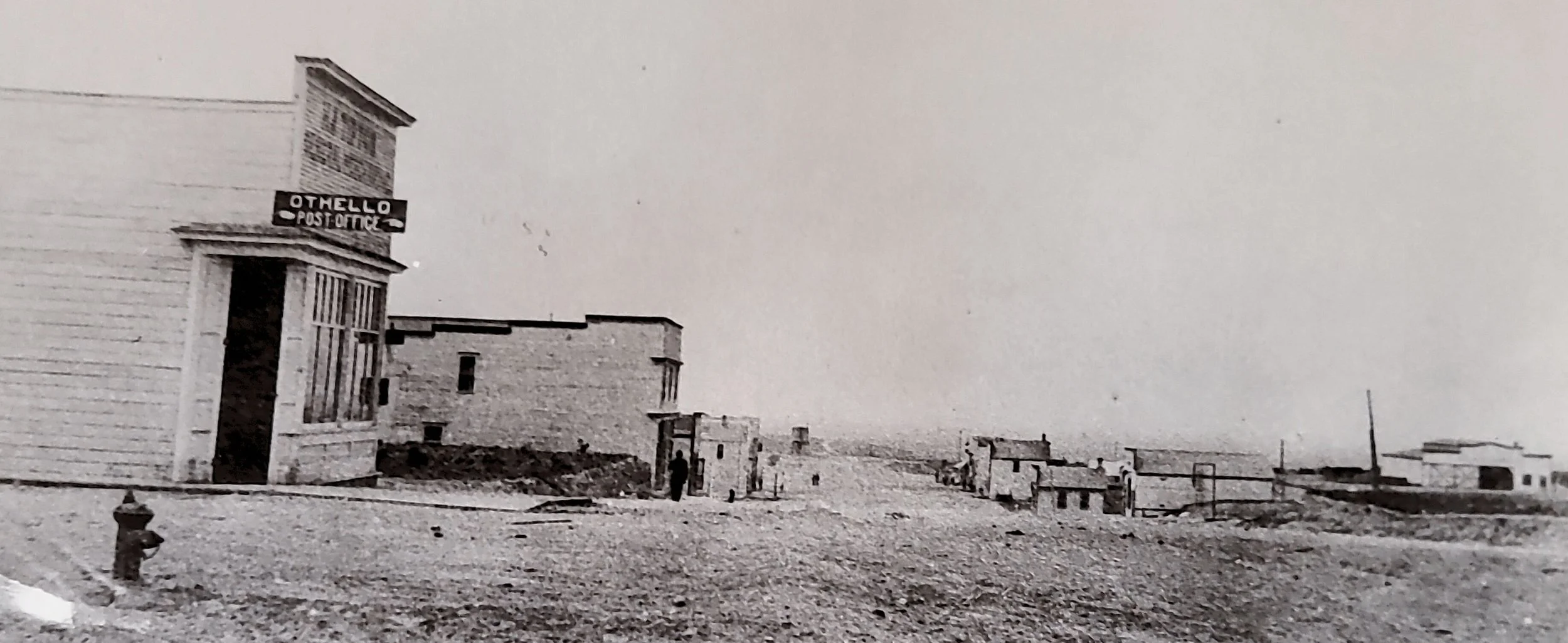 Old black and white photo of a deserted town with a shop labeled 'Othello Post Office' on the left and scattered buildings in the distance.