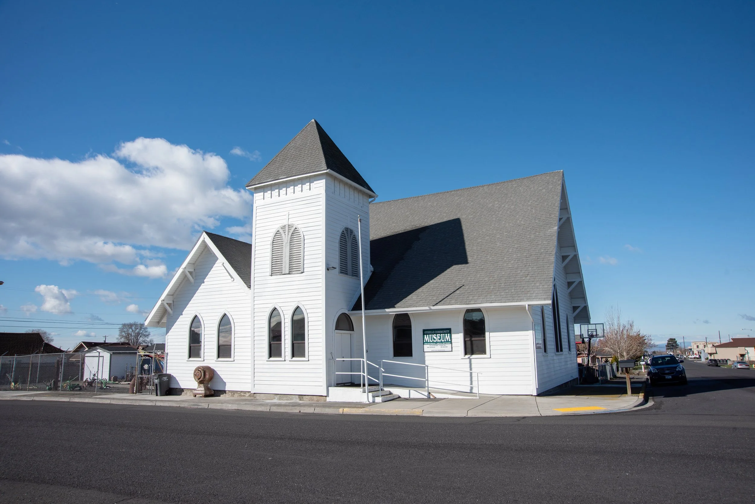 A white wooden church with a steeple and arched windows, located in a small town with a parking lot, a basketball hoop, and nearby residential buildings under a blue sky with some clouds.