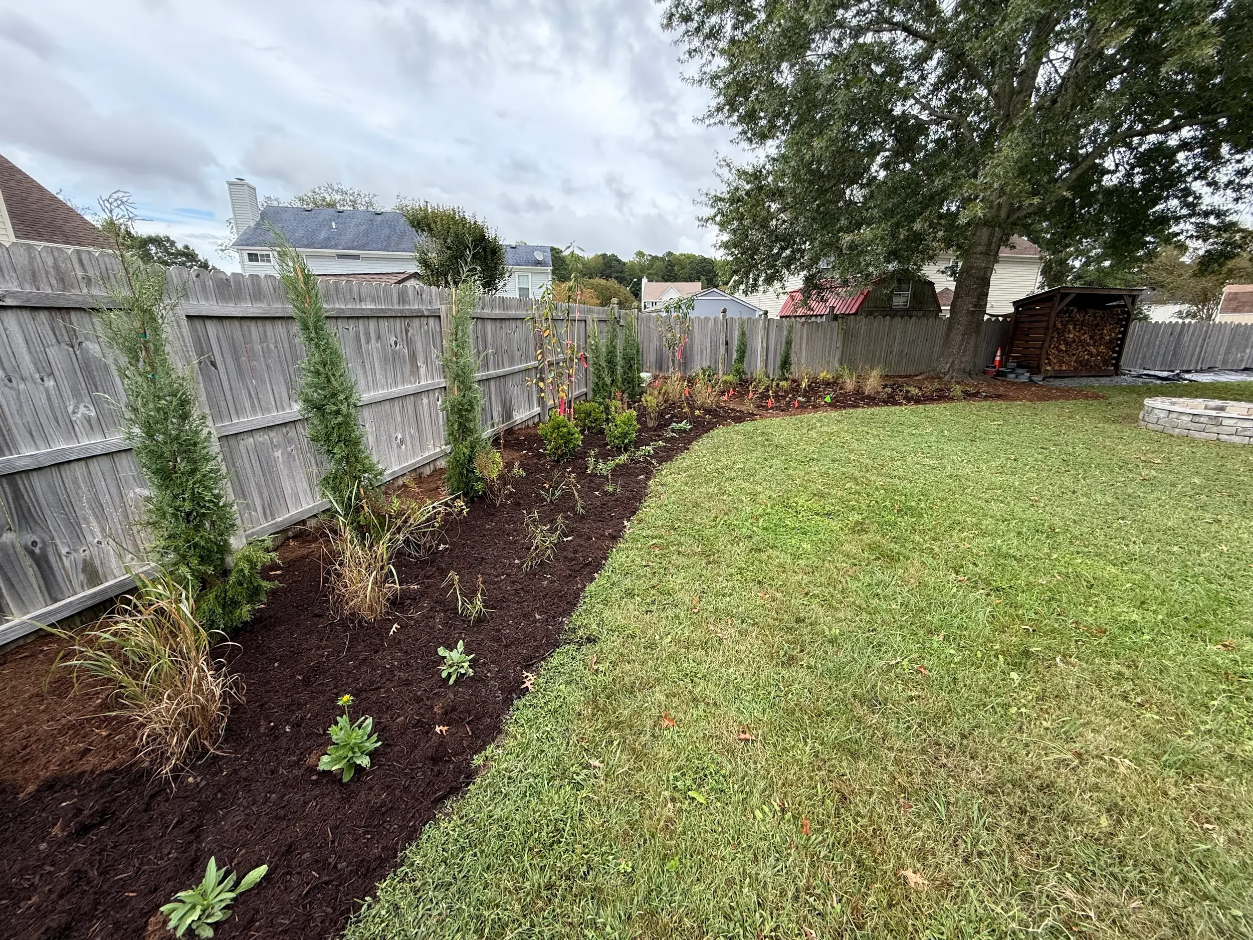 A backyard with a freshly planted flower bed along a wooden fence, bordered by grass, with trees and neighboring houses visible in the background under a cloudy sky.