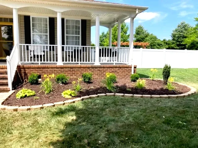 Front yard landscape with freshly planted flower bed bordered by bricks, surrounding a white porch with columns, in front of a house with beige siding and large windows.