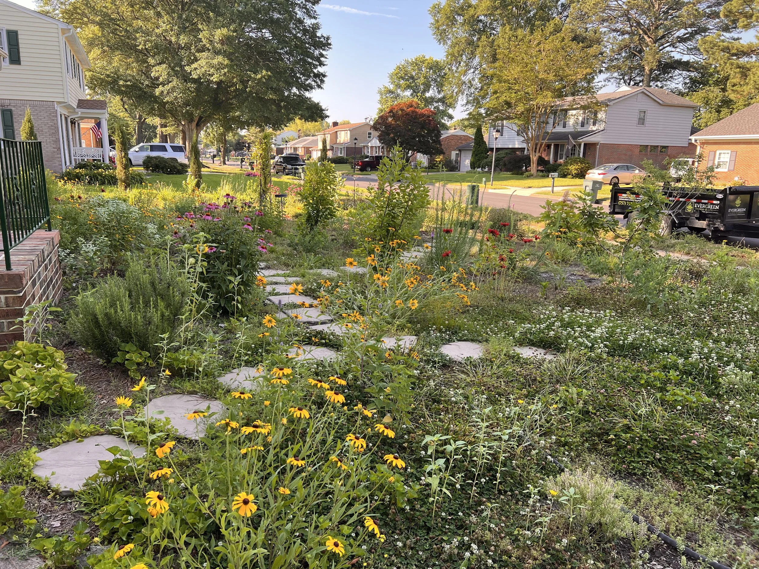 A front yard with a flower garden, pathway, and residential houses in a neighborhood, under a clear blue sky.