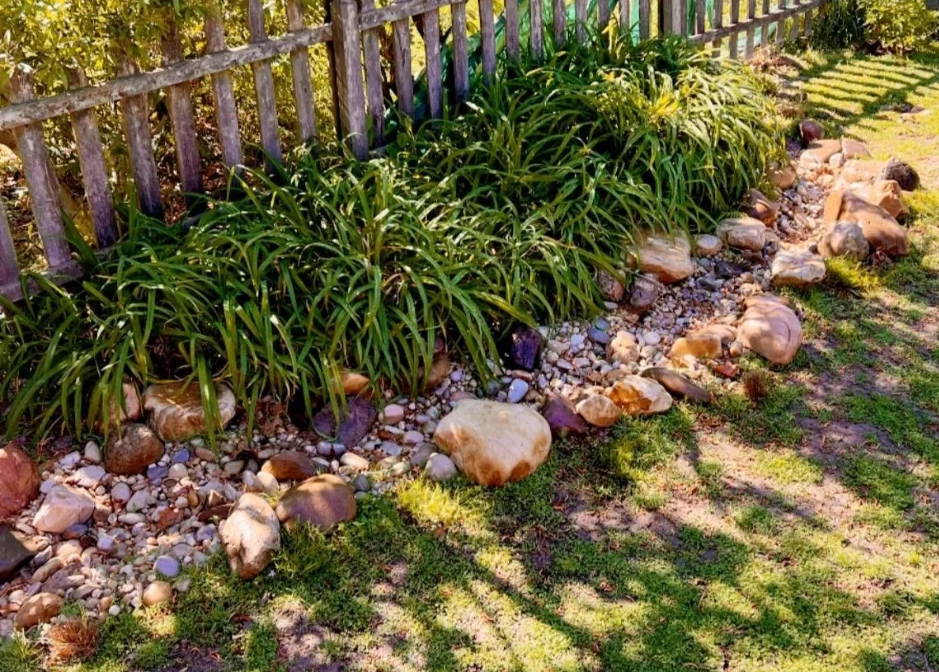 Evergreen Eco-Scapes Project; Ecological Landscaping; A garden bed with green plants along a wooden fence, surrounded by rocks and pebbles, with grass and shadowed patches on the ground.