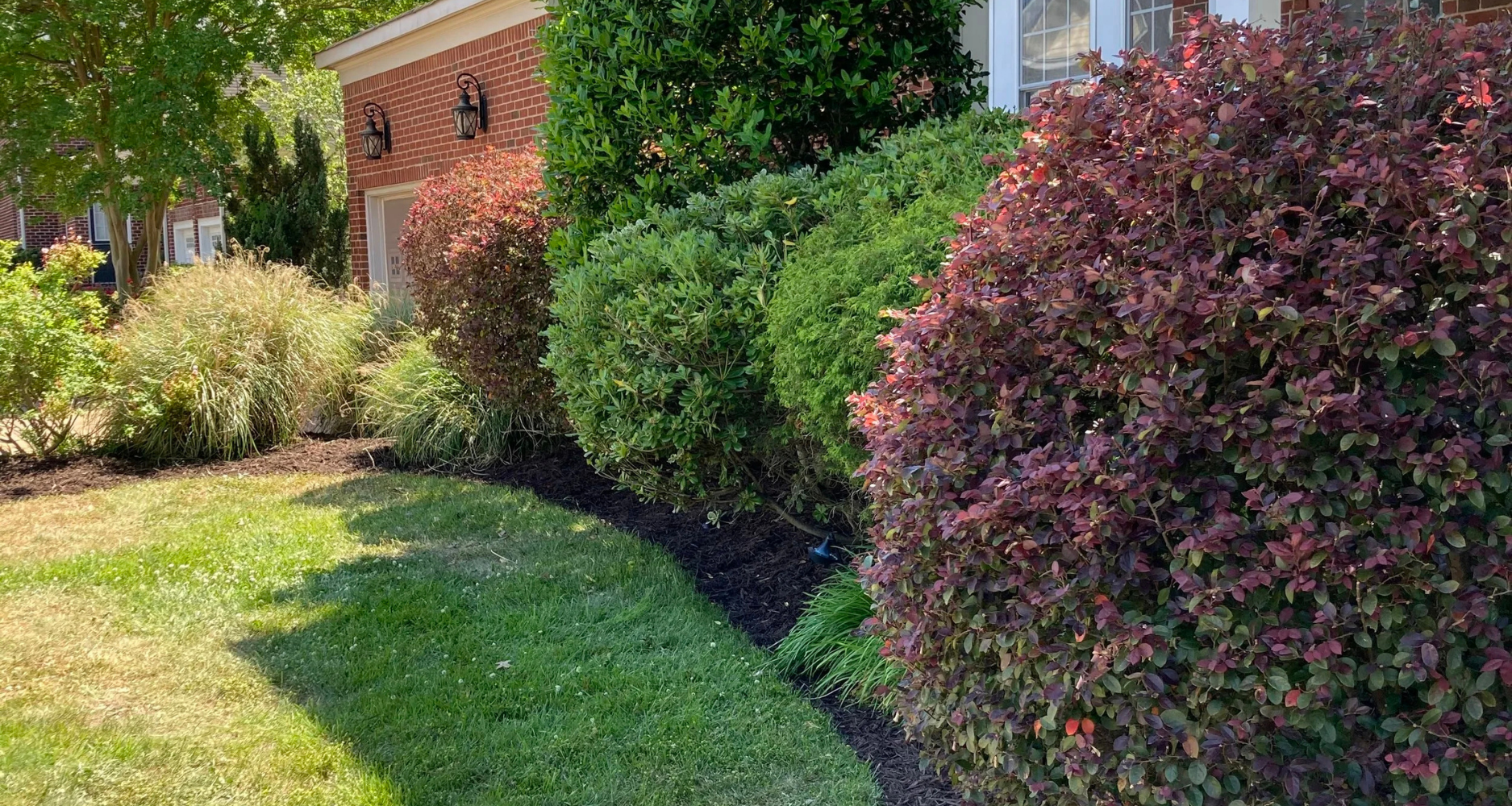 A well-maintained front yard with various bushes, flowers, and a green lawn in front of a brick house with black lantern-style outdoor lights.