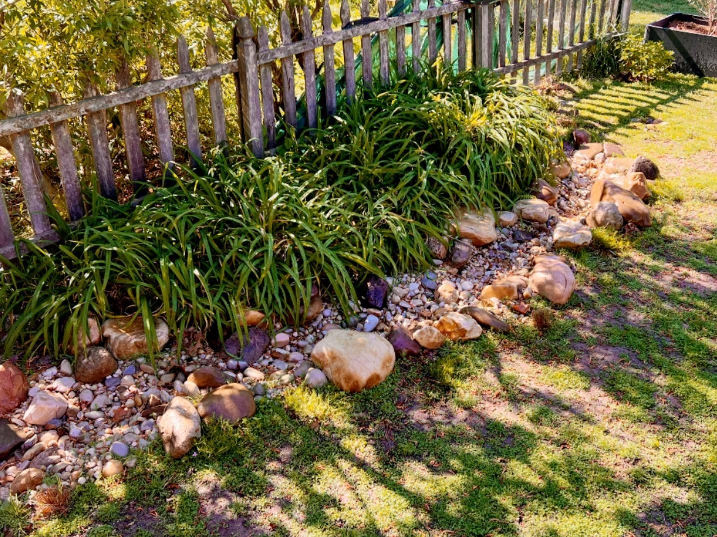 A garden bed with green plants, a wooden fence, rocks, and pebbles, with grass and sunlight casting shadows.