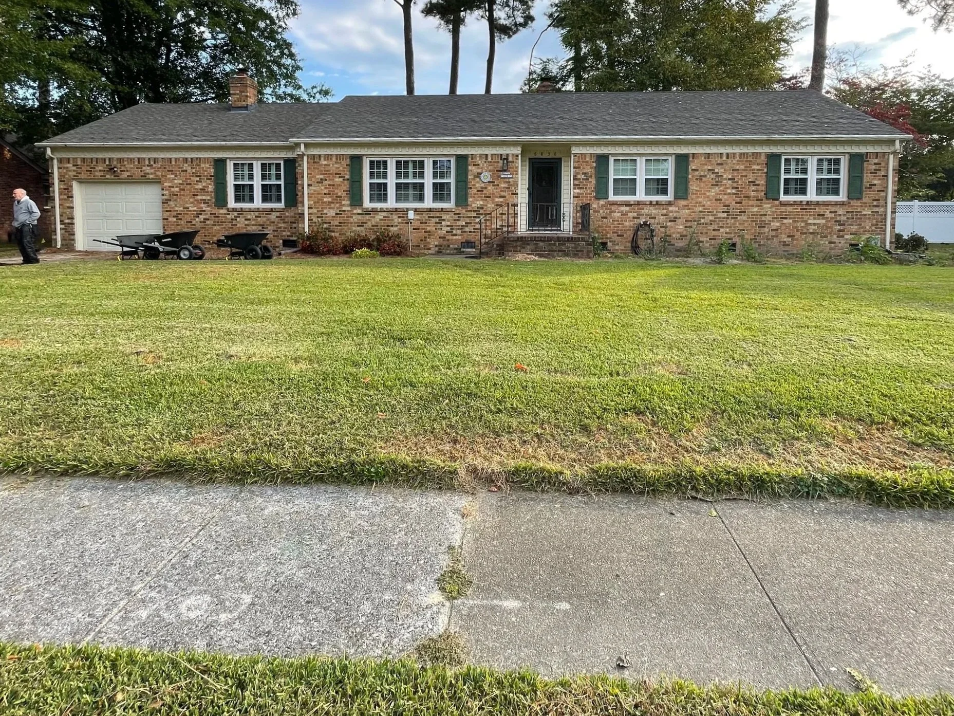 A single-story brick house with green shutters, a small front porch with stairs, and a lawn with some gardening tools and plants. A man stands near the left side of the house, and there are trees in the background.