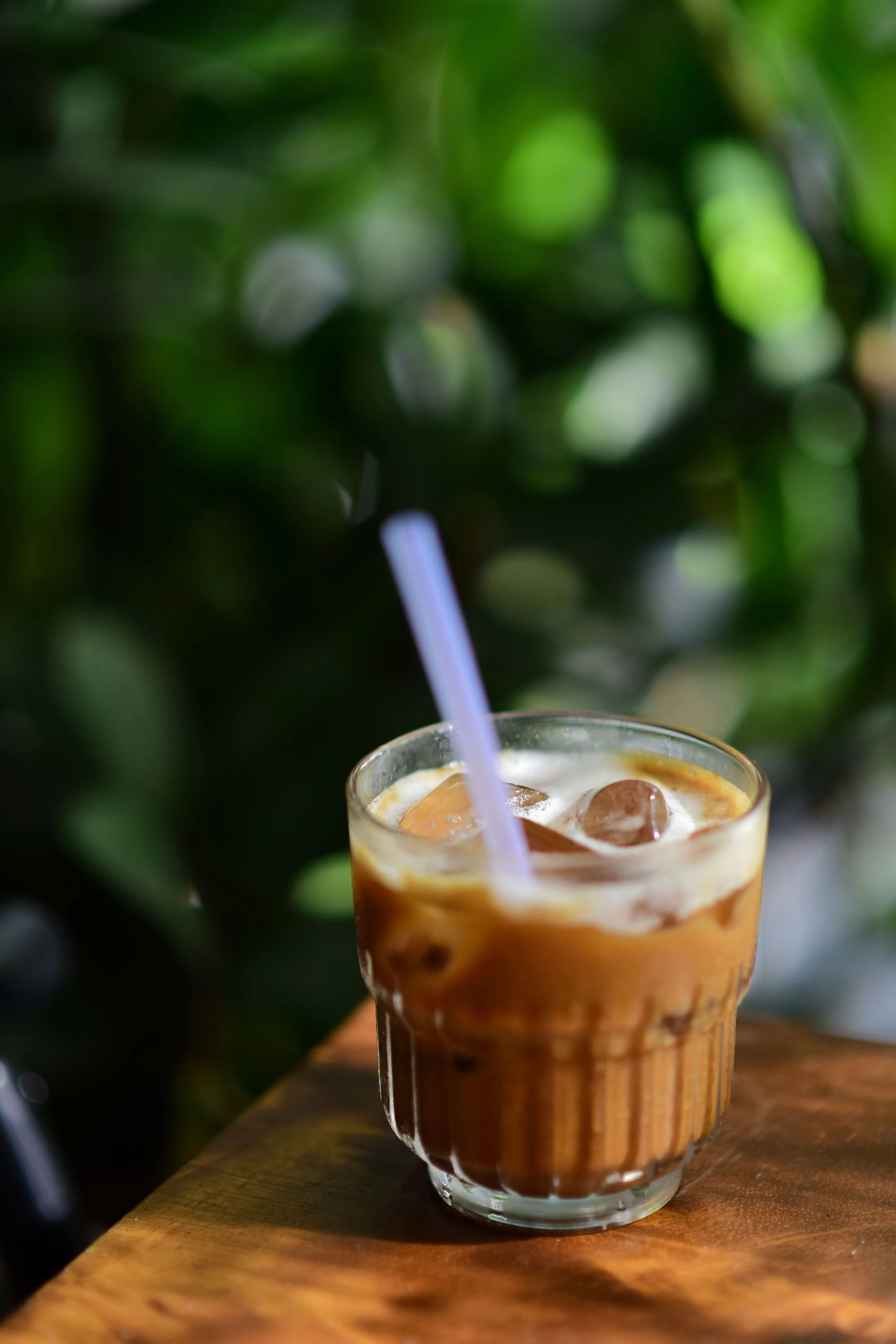 A glass of iced caramel coffee with a straw on a wooden surface, with blurred greenery in the background.