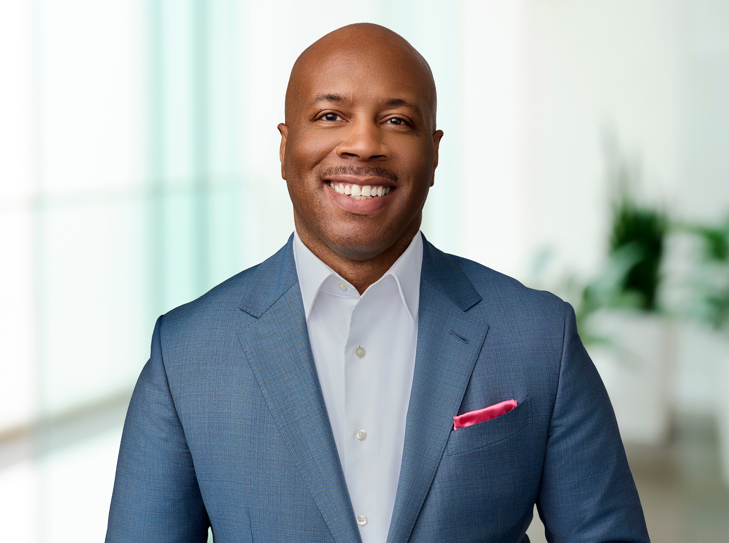 A portrait of a smiling Black man in a dark blue suit with a white shirt and a pink pocket square.