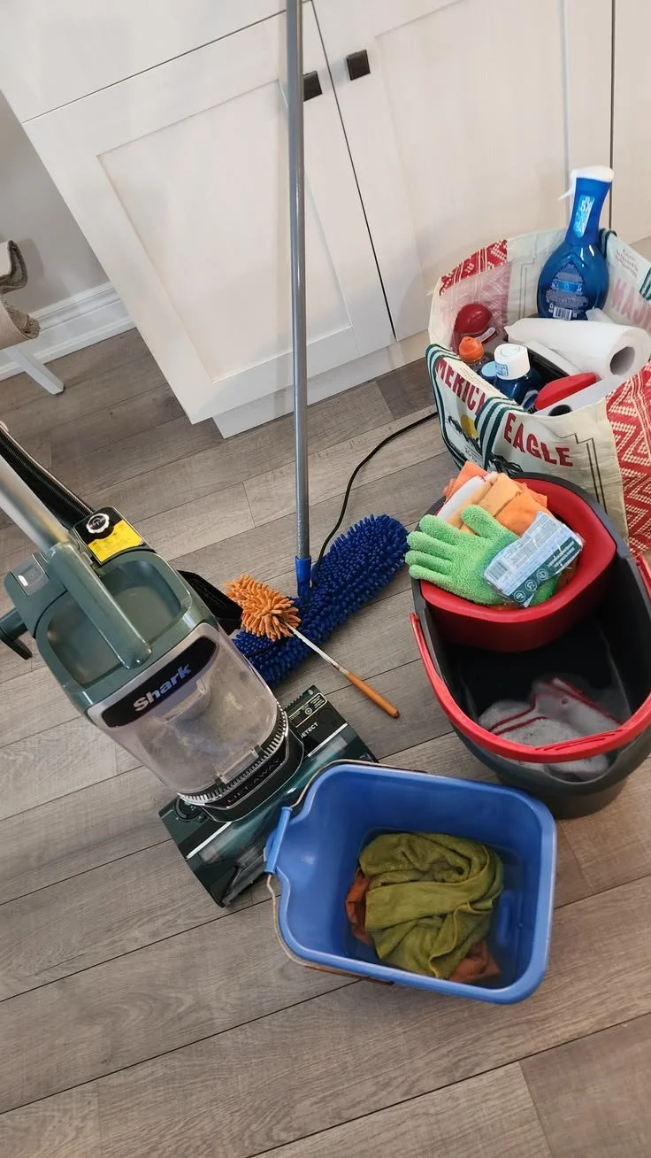 Cleaning supplies including a Shark vacuum, mop, cleaning cloths, gloves, and various cleaning products arranged on a hardwood floor.