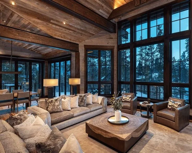 Luxury living room with large windows, wooden ceiling, beige sofas, armchairs with patterned pillows, a wooden coffee table with a flower arrangement, side tables, and lamps, overlooking a snowy outdoor scene.