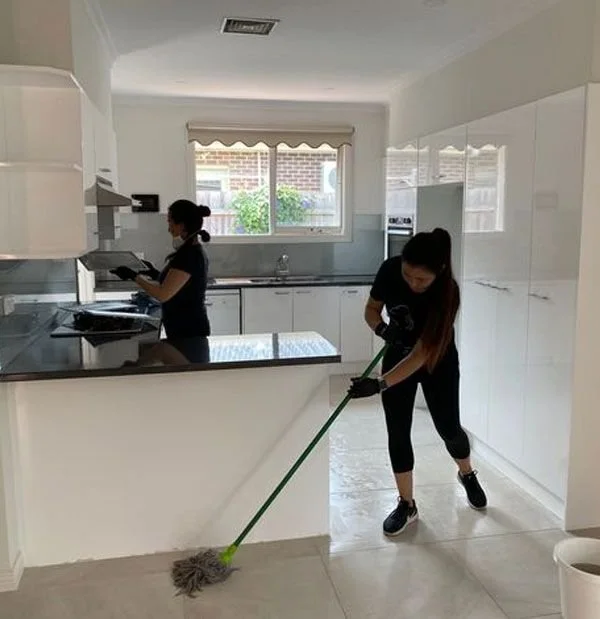 Two women cleaning a bright kitchen, one is sweeping the floor while the other is reading a recipe or instructions.