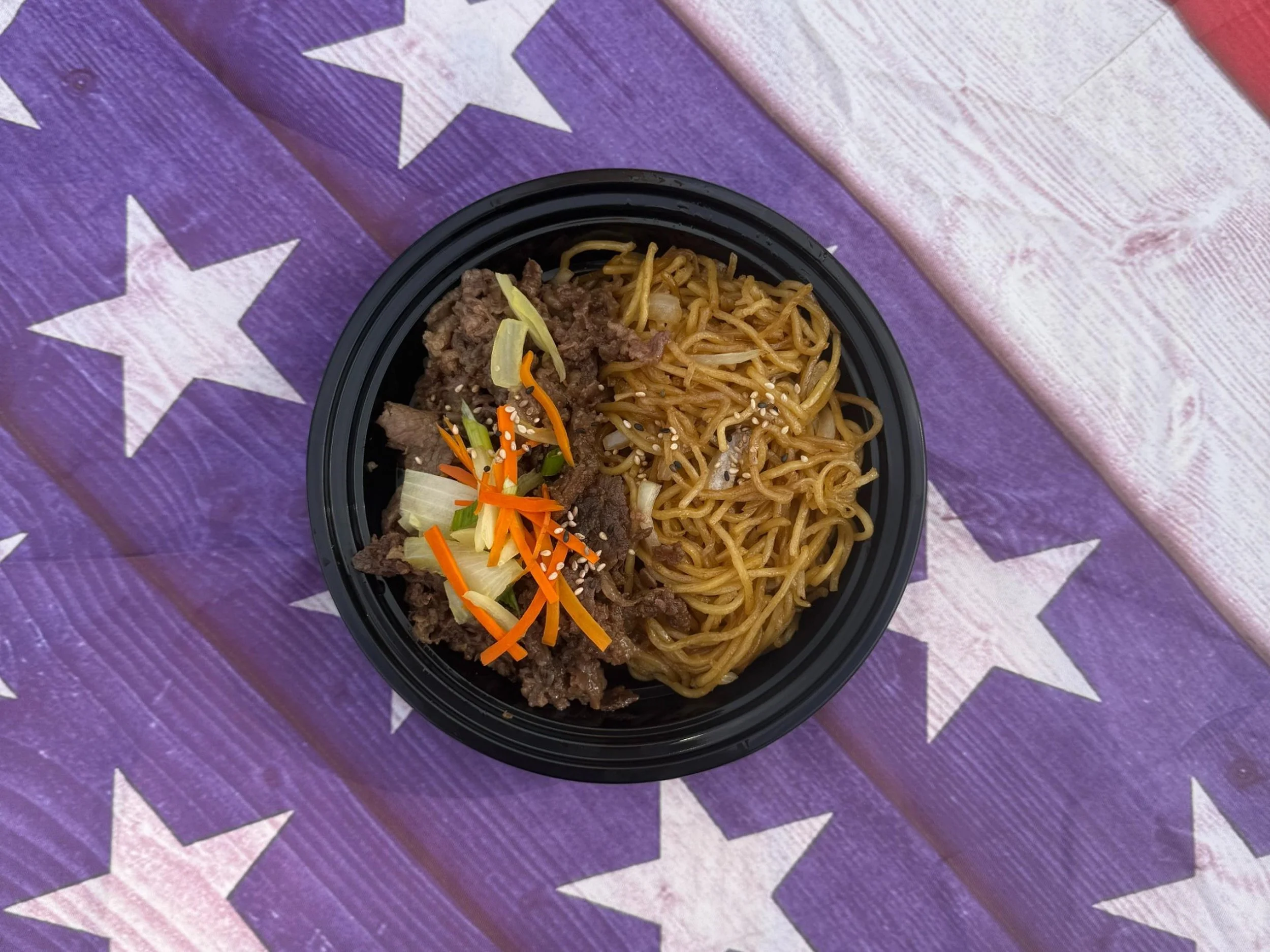 A takeout bowl of Asian-style noodles with ground beef, shredded carrots, and cabbage garnished with sesame seeds, placed on a decorated purple star-patterned tablecloth.