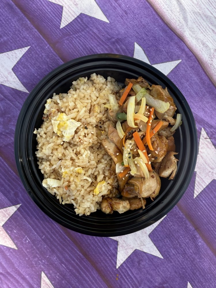 A black bowl contains fried rice with bits of scrambled egg and a portion of stir-fried chicken with vegetables, including carrots, onions, and scallions, placed on a purple tablecloth with white stars.