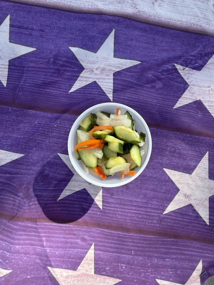 A small bowl of sliced cucumbers, carrots, and onions on a purple tablecloth with white star patterns.