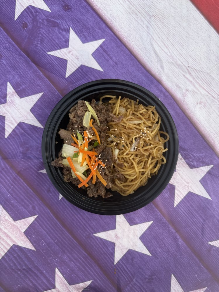 A black bowl of stir-fry noodles with beef, chopped vegetables, and sesame seeds on top, placed on a purple wooden surface with white stars.