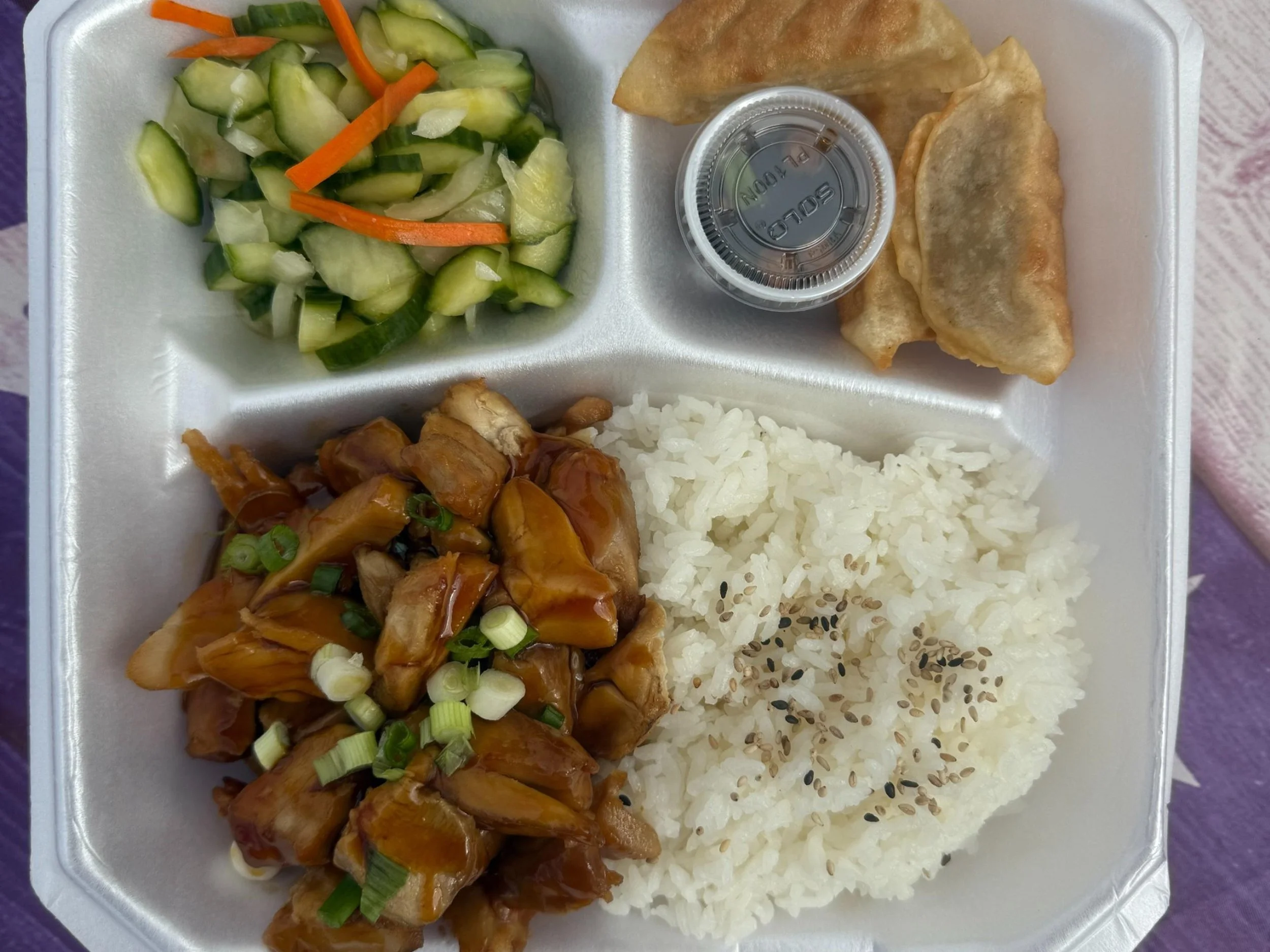A foam container with Asian food including cucumber and carrot salad, a container of dipping sauce, two fried dumplings, a serving of cooked chicken in sauce garnished with chopped green onions, a portion of white rice with black sesame seeds, and a side of stir-fried vegetables.