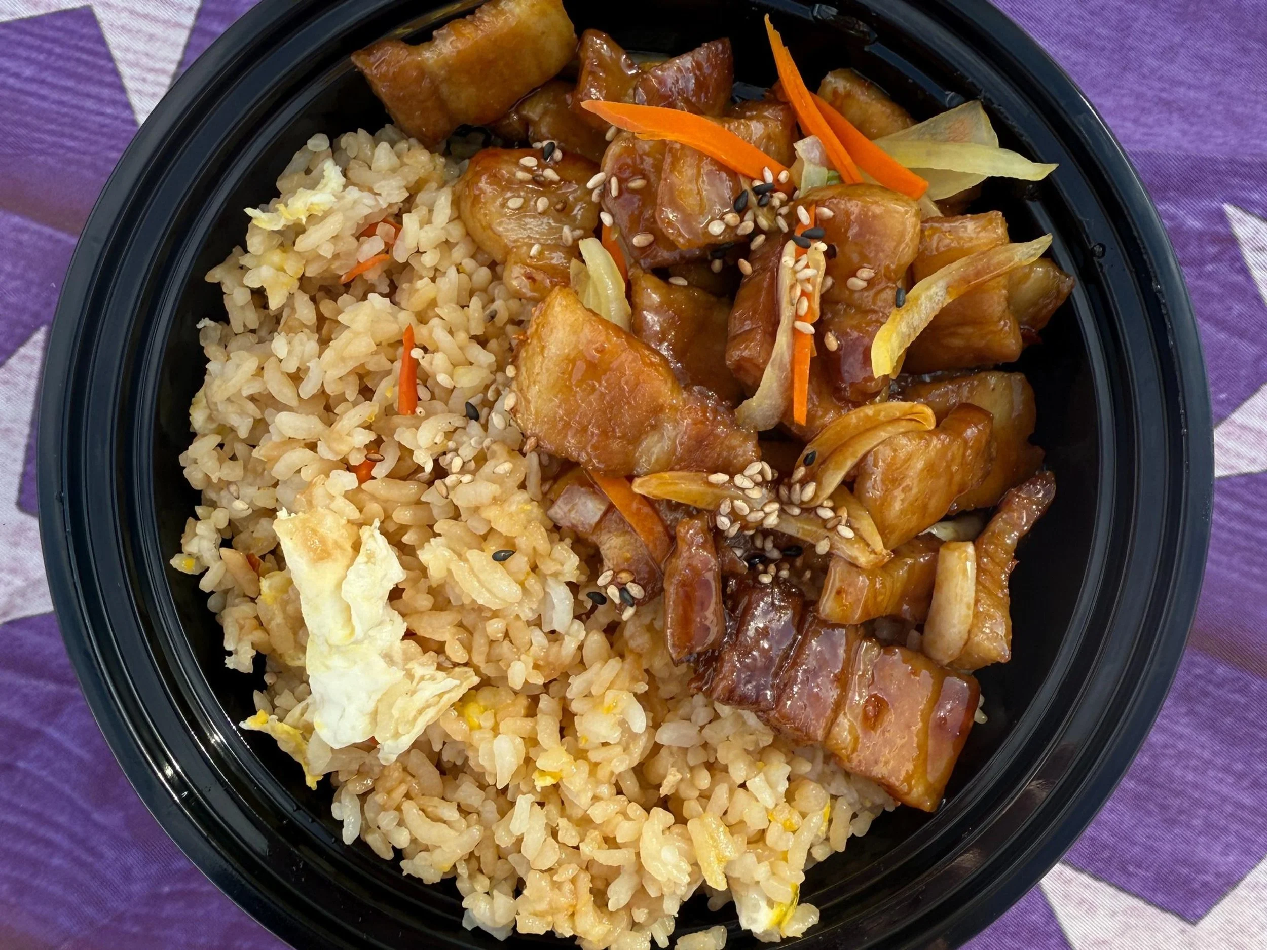 Close-up of a black container filled with fried rice and sweet and sour chicken topped with sesame seeds and shredded carrots, set on a purple tablecloth.
