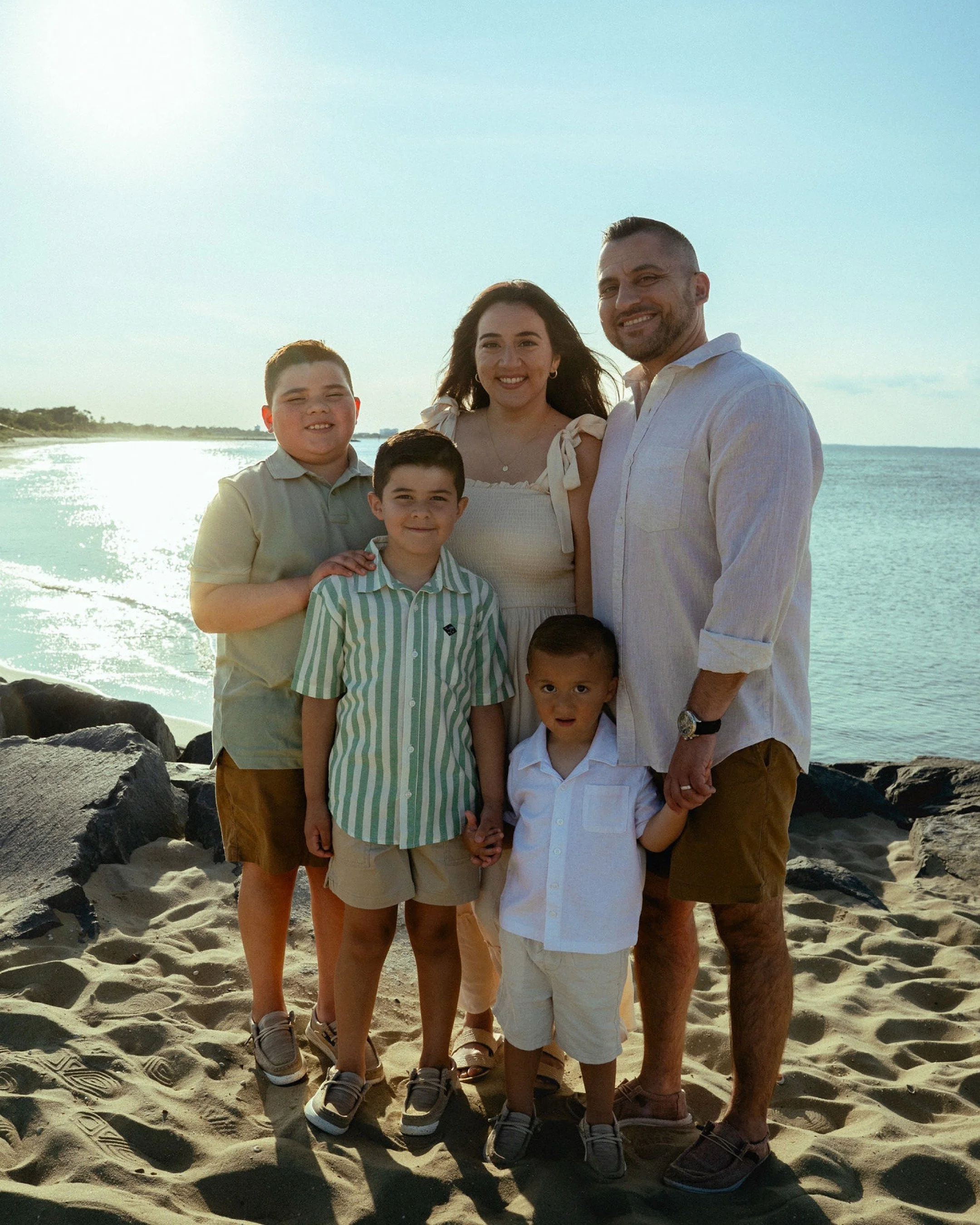 A family standing on a sandy beach near the ocean, smiling at the camera during sunset.