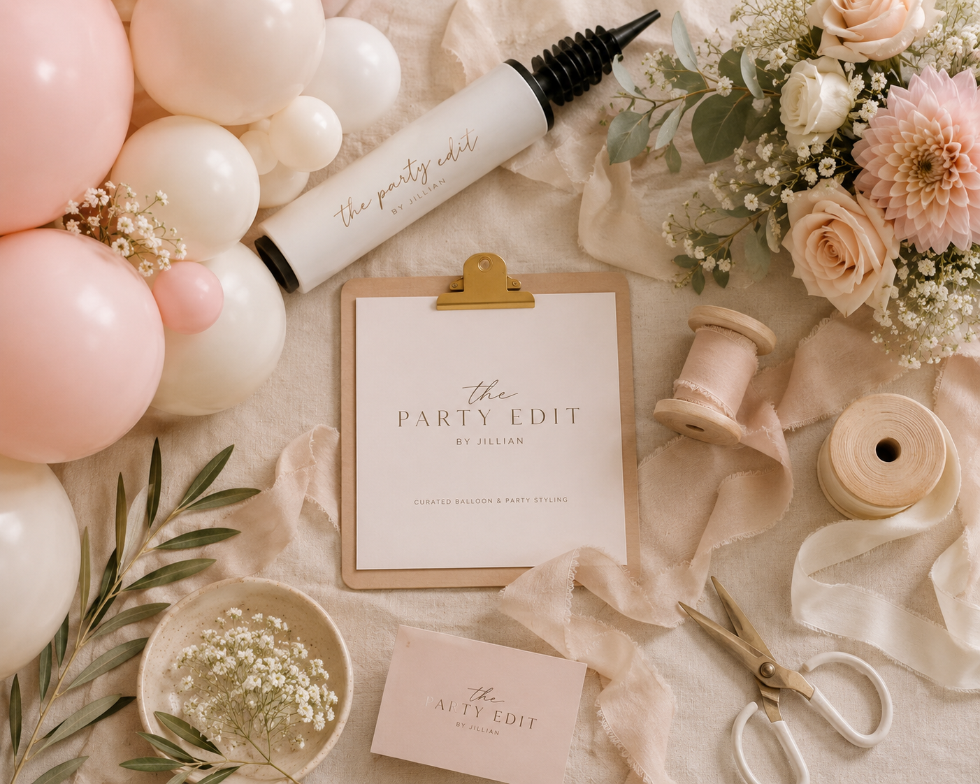 Arrangement of pink and white balloons, a bouquet of pink and white roses and other flowers, a balloon decorating kit with a piping gun labeled ‘the party edit by Jillian,’ a clipboard with a card titled ‘the PARTY EDIT by Jillian,’ a small bowl of baby's breath flowers, ribbon spools, and a pair of scissors, all set on a beige fabric background for party decoration planning.