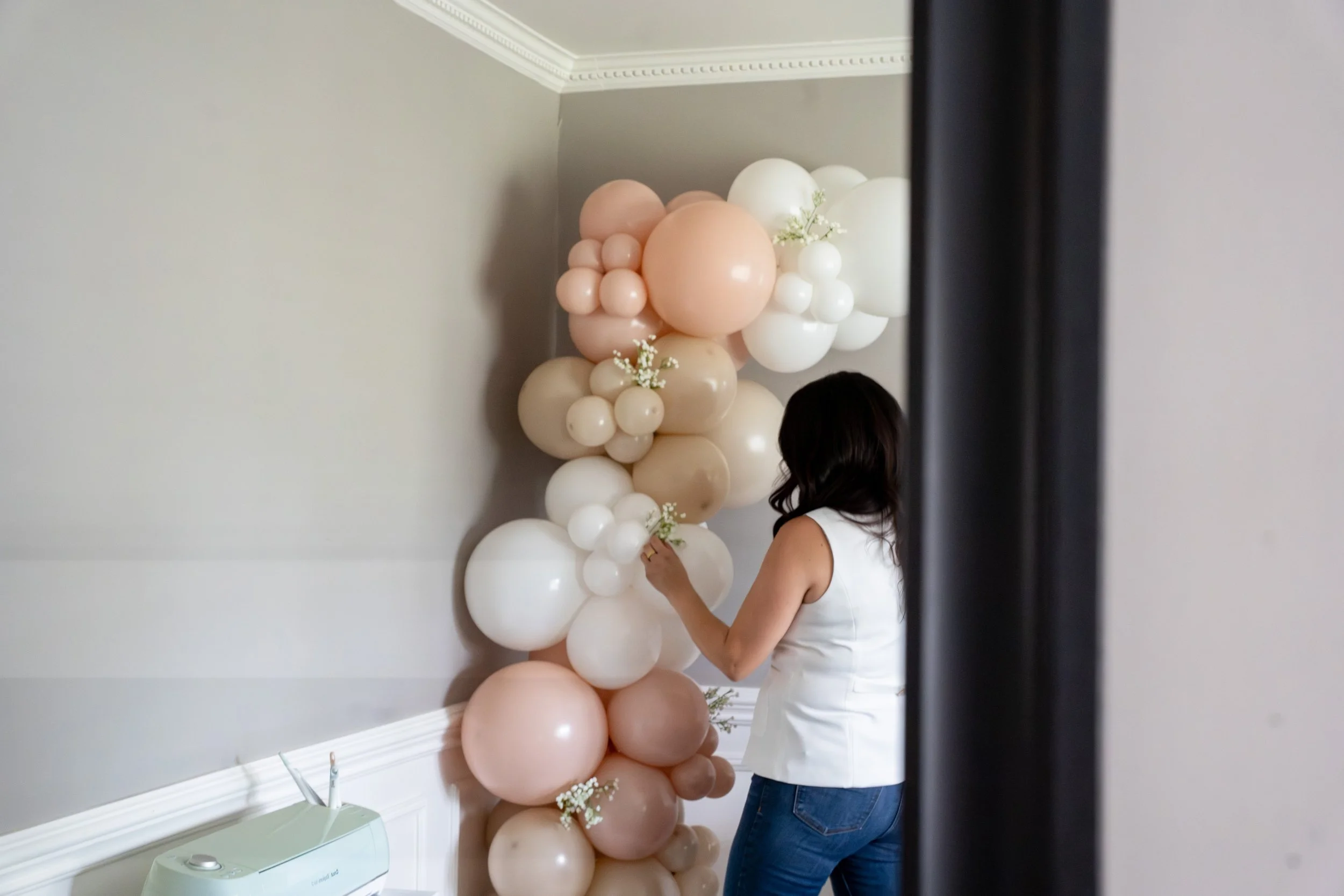 A woman placing balloons on a balloon arch in a room with light gray walls and crown molding.