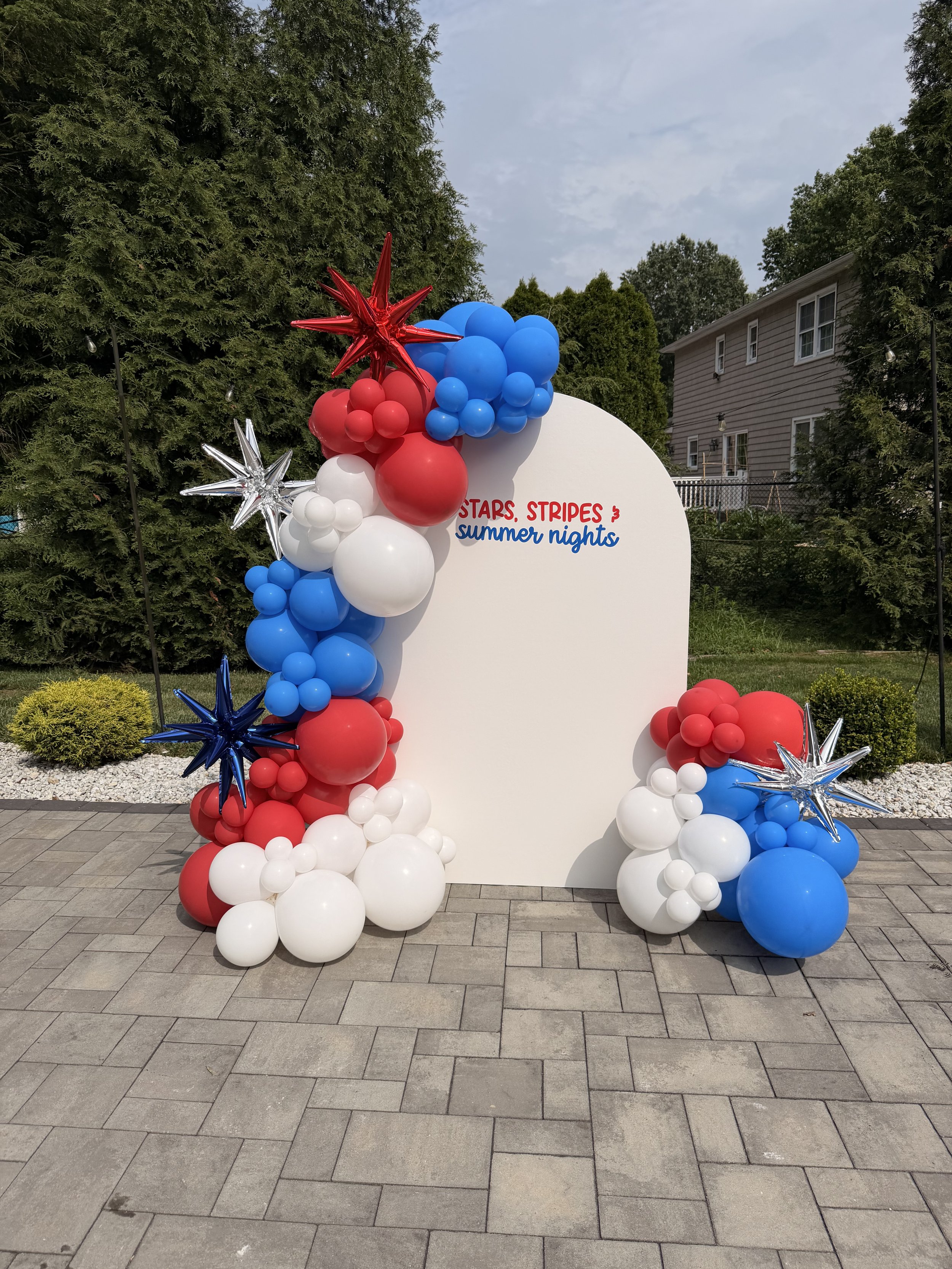 Decoration with red, white, and blue balloons and star-shaped foil balloons arranged around a white sign with red and blue text that reads 'Stars, Stripes & Summer Nights' outdoors on a paved area with trees and a house in the background.