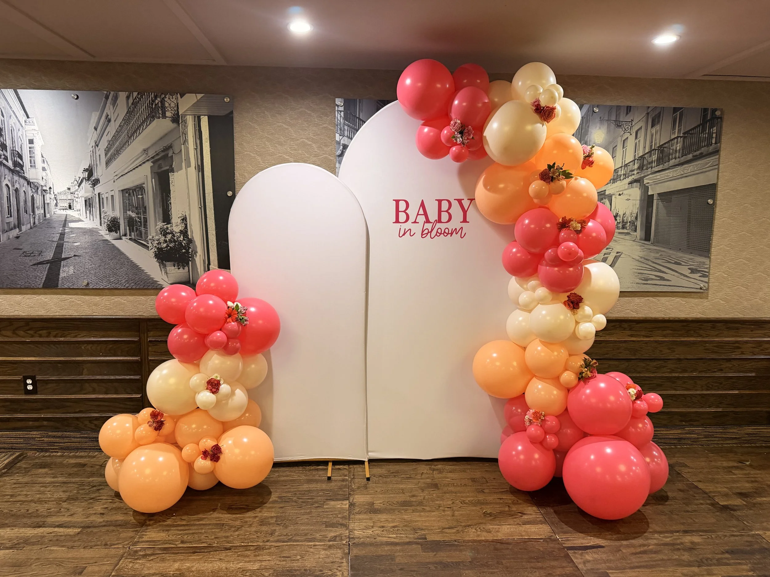 Decorative backdrop with pink, peach, and cream balloons arranged around a white panel that says 'BABY in bloom' with small flowers attached. Black and white mural of a European street scene in the background.
