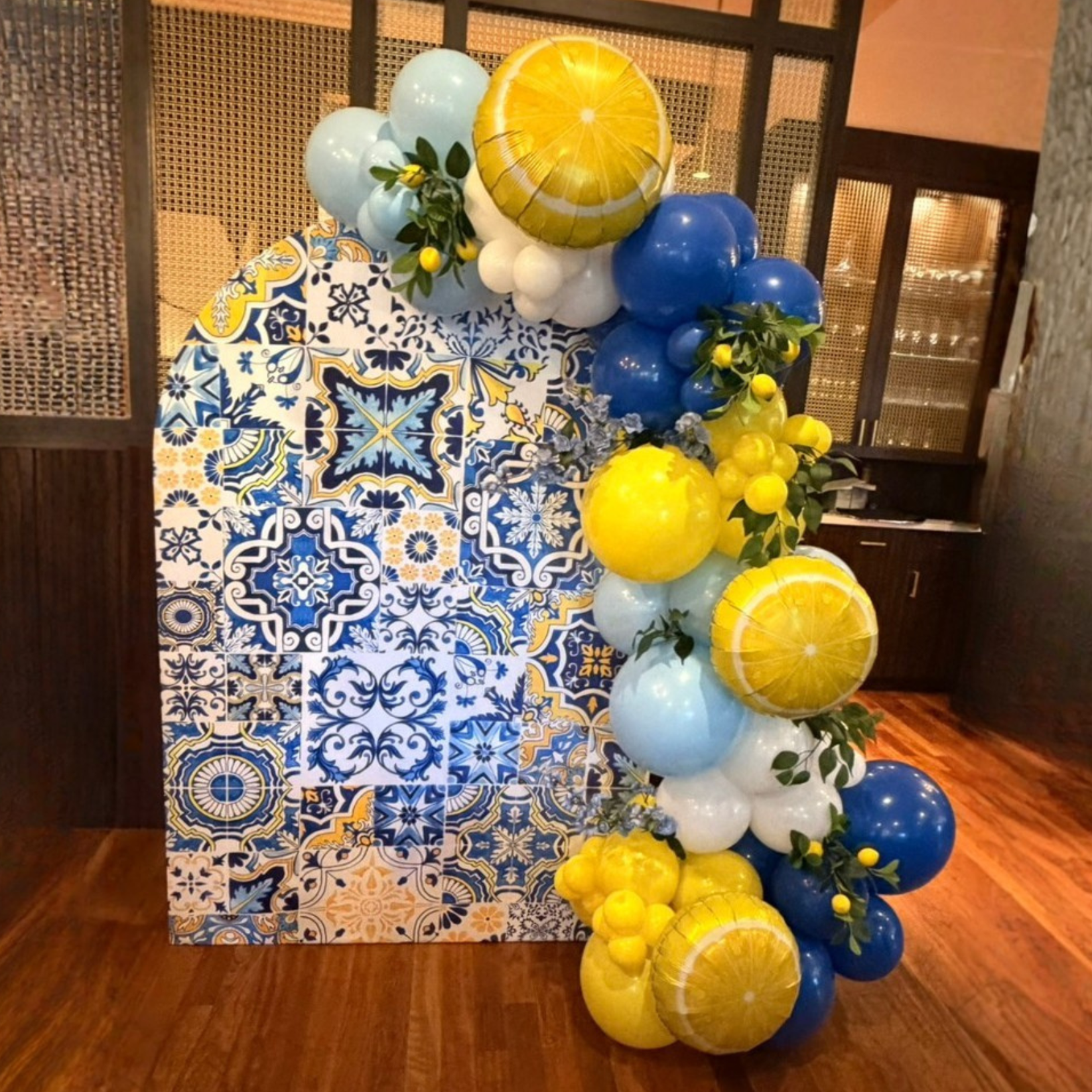 A decorative display of blue, yellow, and white balloons arranged on a table with a patterned cloth, paired with green foliage and yellow lemons, in a warm wooden-floored room.