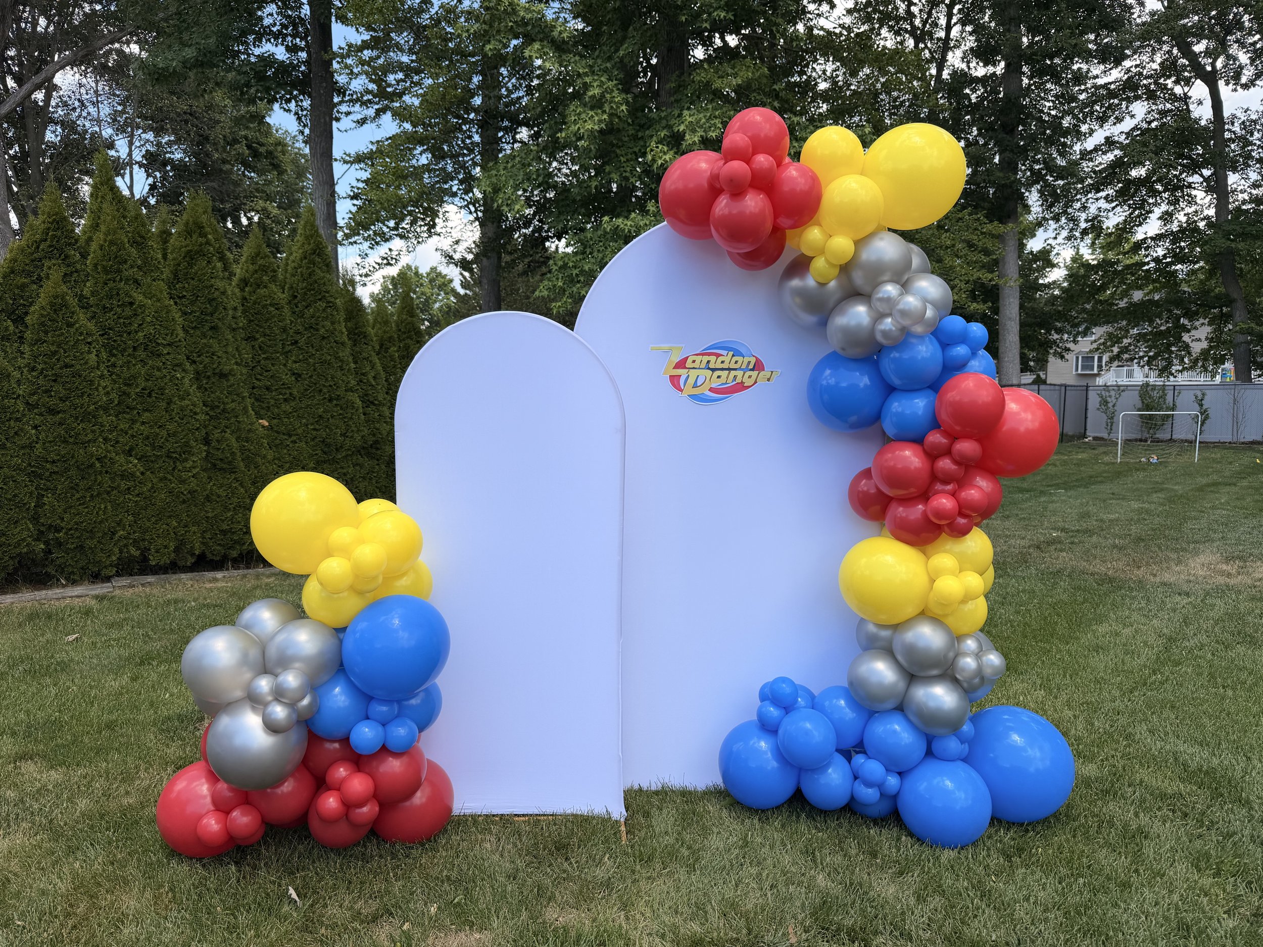 A colorful balloon arch with red, yellow, blue, and silver balloons surrounds a white backdrop with the Landon Dancer logo, set outdoors among trees and grass.