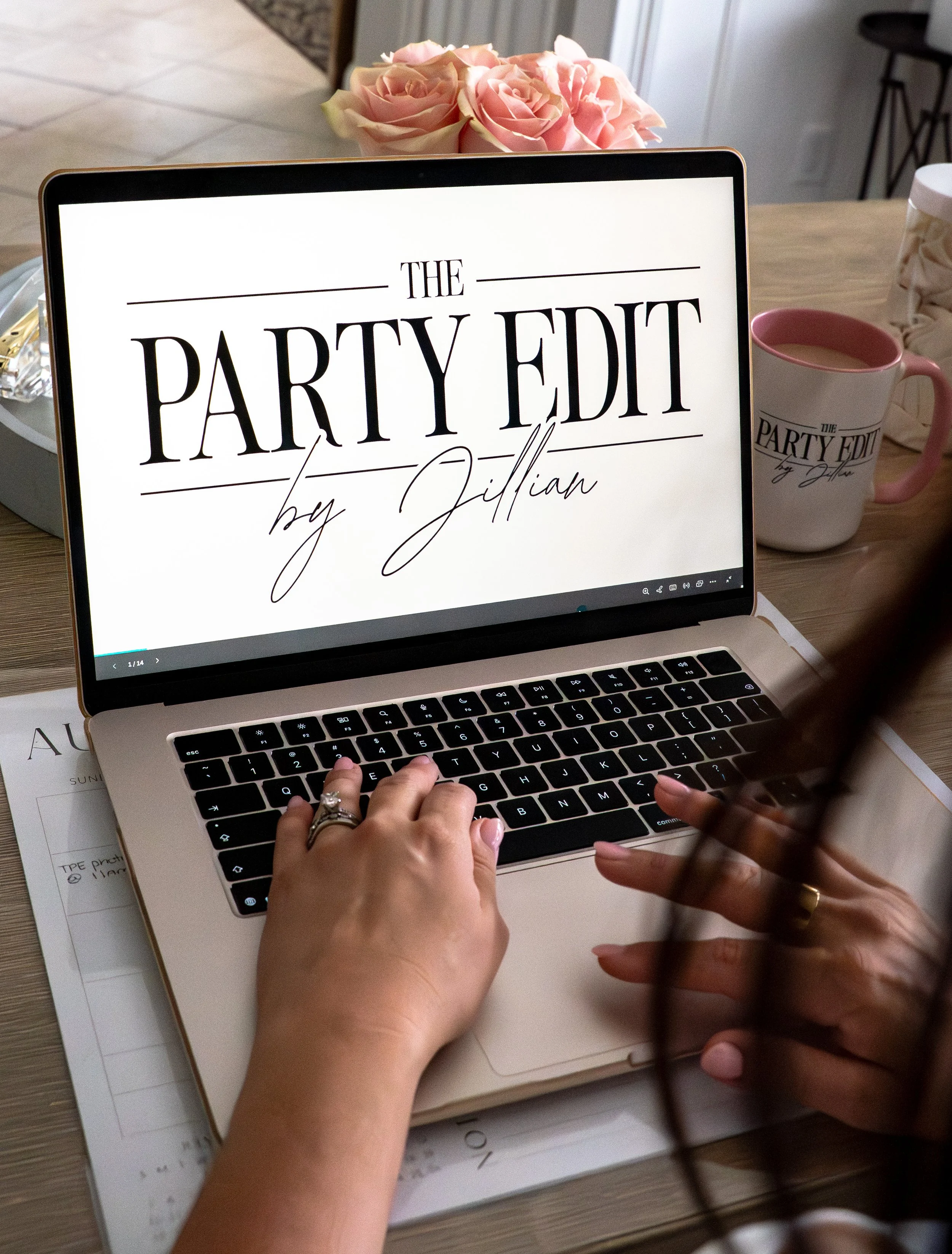 A laptop on a table displays the text 'The Party Edit by Jillian'. A person's hands are typing on the keyboard. Behind the laptop, there is a mug with the same text, a bouquet of pink roses, and some papers.