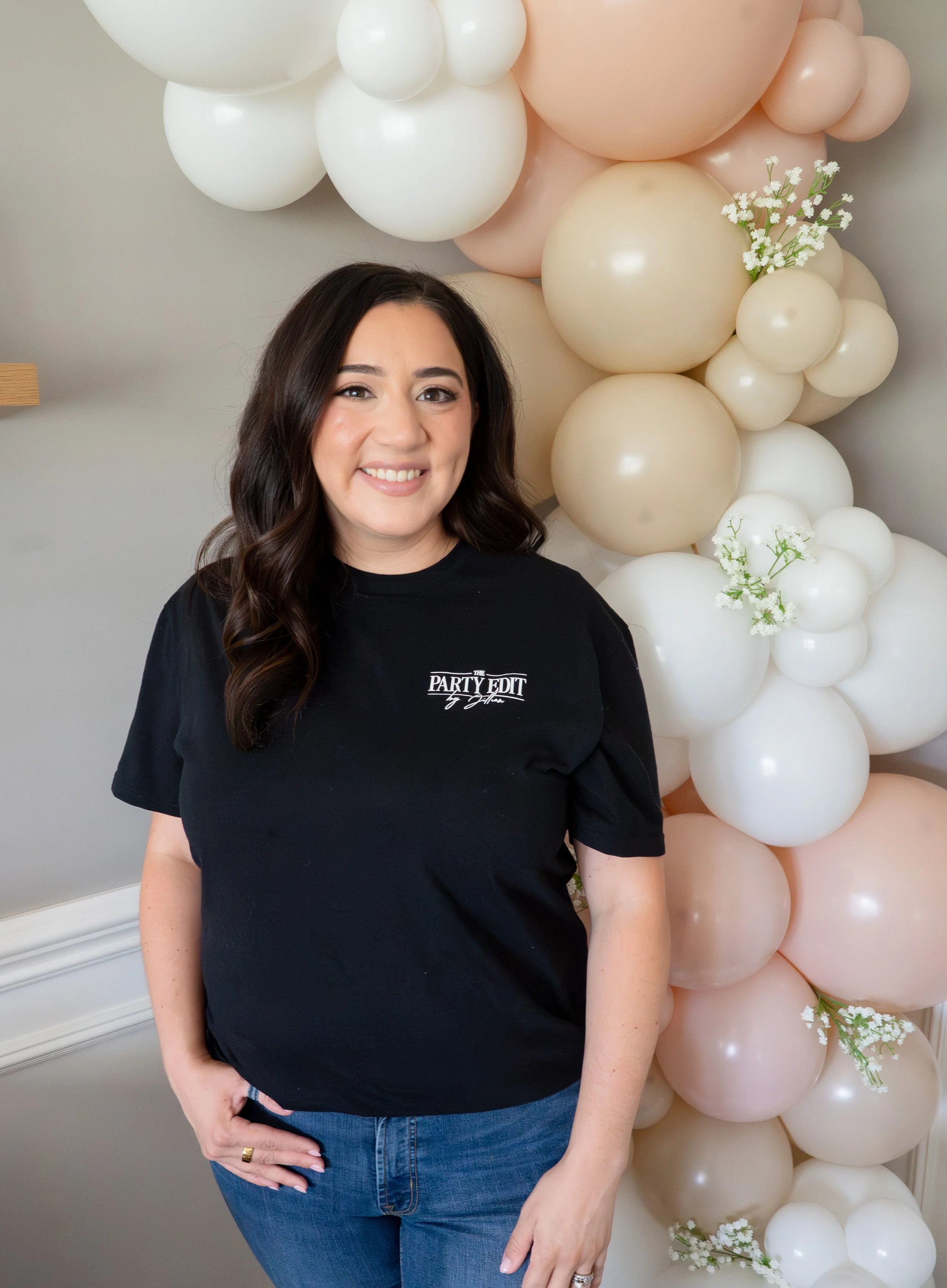 A woman with dark hair smiling, wearing a black shirt and blue jeans, standing beside a balloon display with pink, cream, and white balloons and some small white flowers near the balloons.