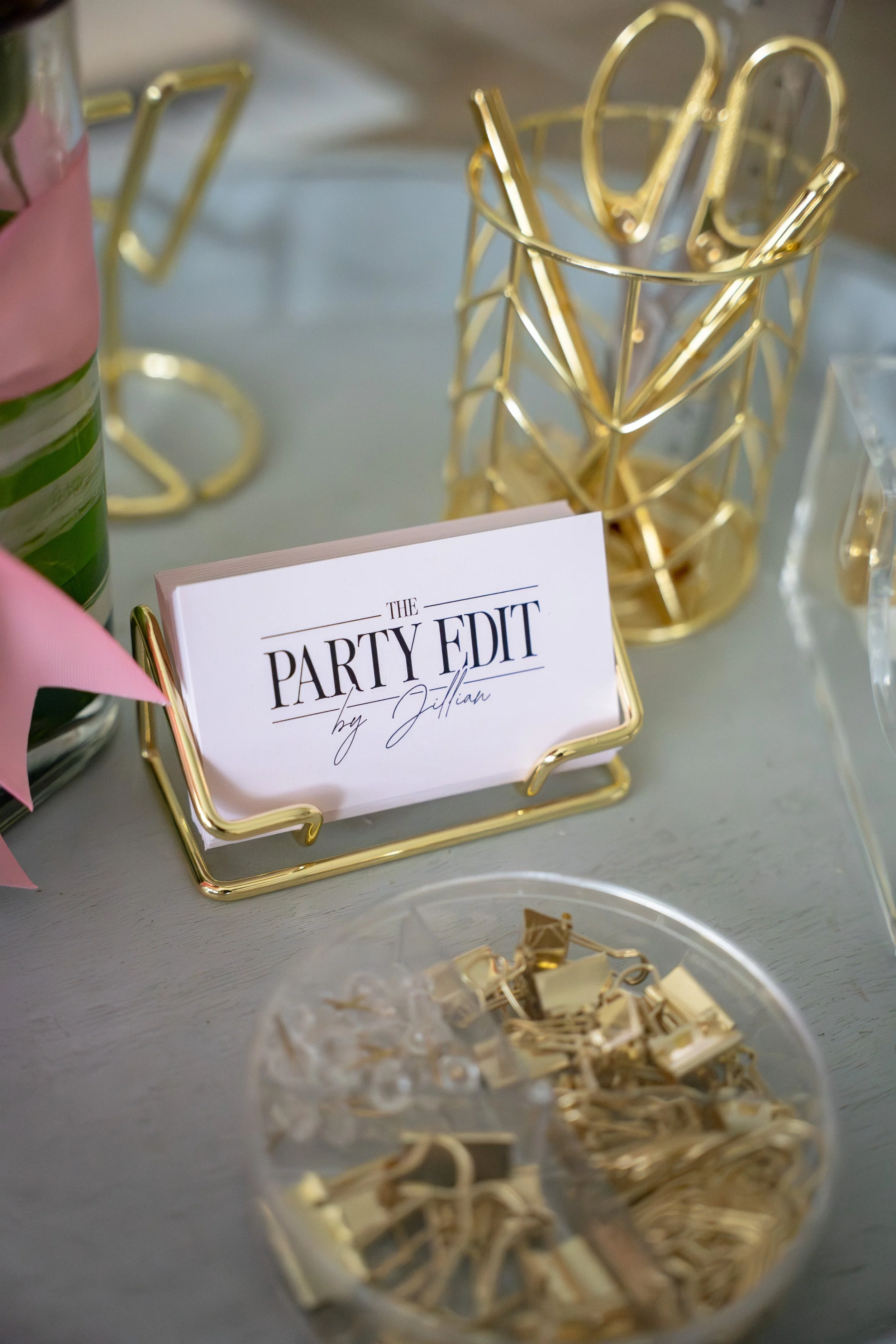 Gold wire basket holding a card that reads "The Party Edit by Jillian" with decorative gold paper clips in a clear bowl in the foreground, a pink and green plant to the side, and other decorative items on a table.