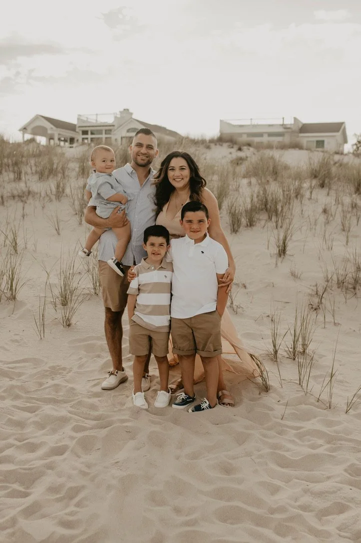 Family of five posing on a sandy beach with dunes and beach houses in the background. The family includes two adults and three children, all smiling.