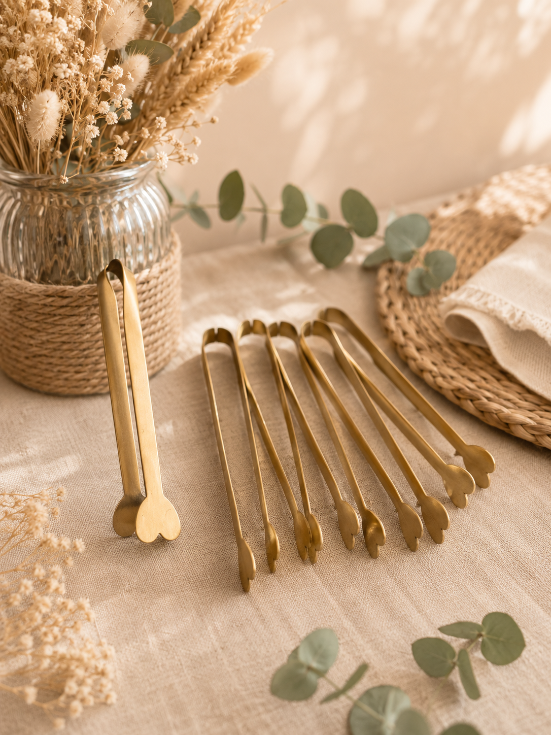 Une table avec des pinces à serviette dorées, une jarre en verre avec des fleurs séchées, et dans le fond un plateau en rotin avec une serviette en tissu, entourés de feuilles d'eucalyptus.