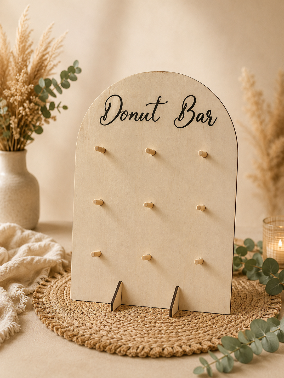 Un tableau en bois clair avec l'inscription « Donut Bar » écrit en cursive noire, posé sur une nappe tressée, décoré de fleurs séchées et d'une petite bougie.