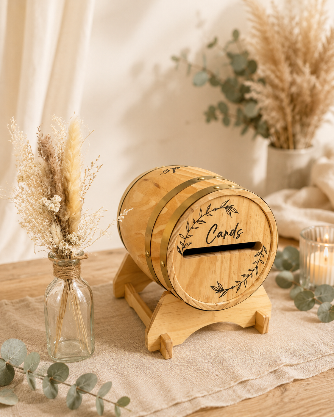 Une boîte en bois en forme de tonneau avec une ouverture pour des cartes, portant l'inscription "Cards" et décorée de motifs de feuilles, posée sur une table en bois avec un vase contenant des fleurs séchées à côté, entourée de décorations naturelles.