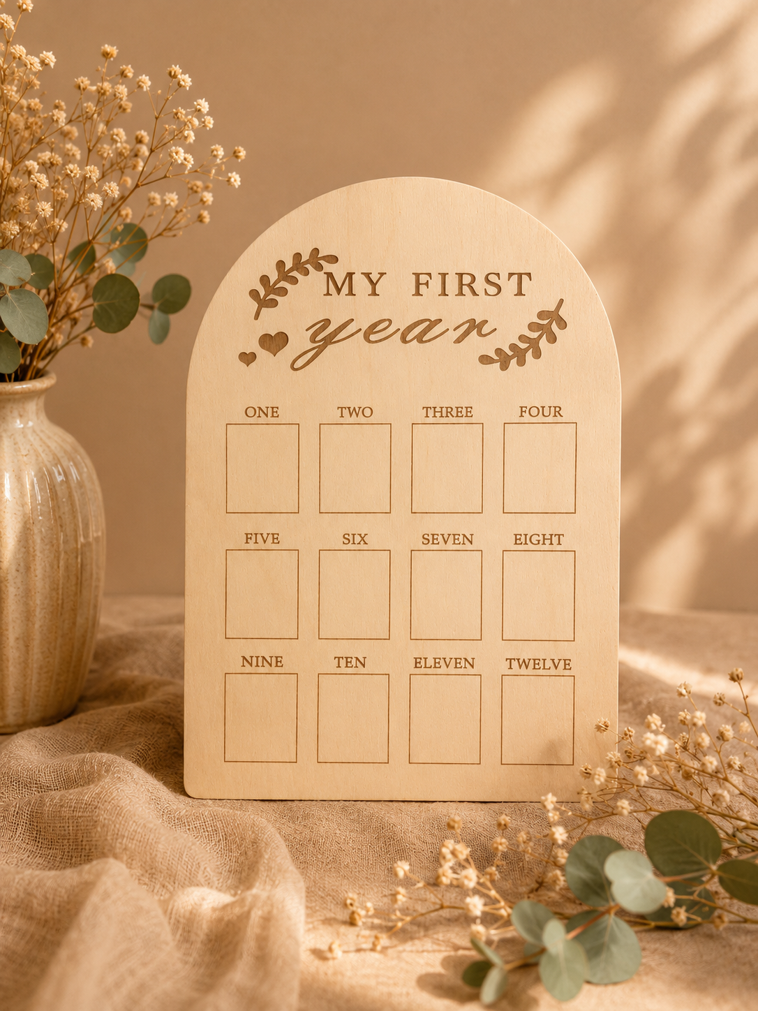 Panneau en bois gravé avec la phrase ''My First Year'' orné de feuilles décoratives, avec des cases pour inscrire les mois en chiffres et en lettres, placé à côté d'un vase de fleurs et de branches d'eucalyptus sur une toile de coton beige.