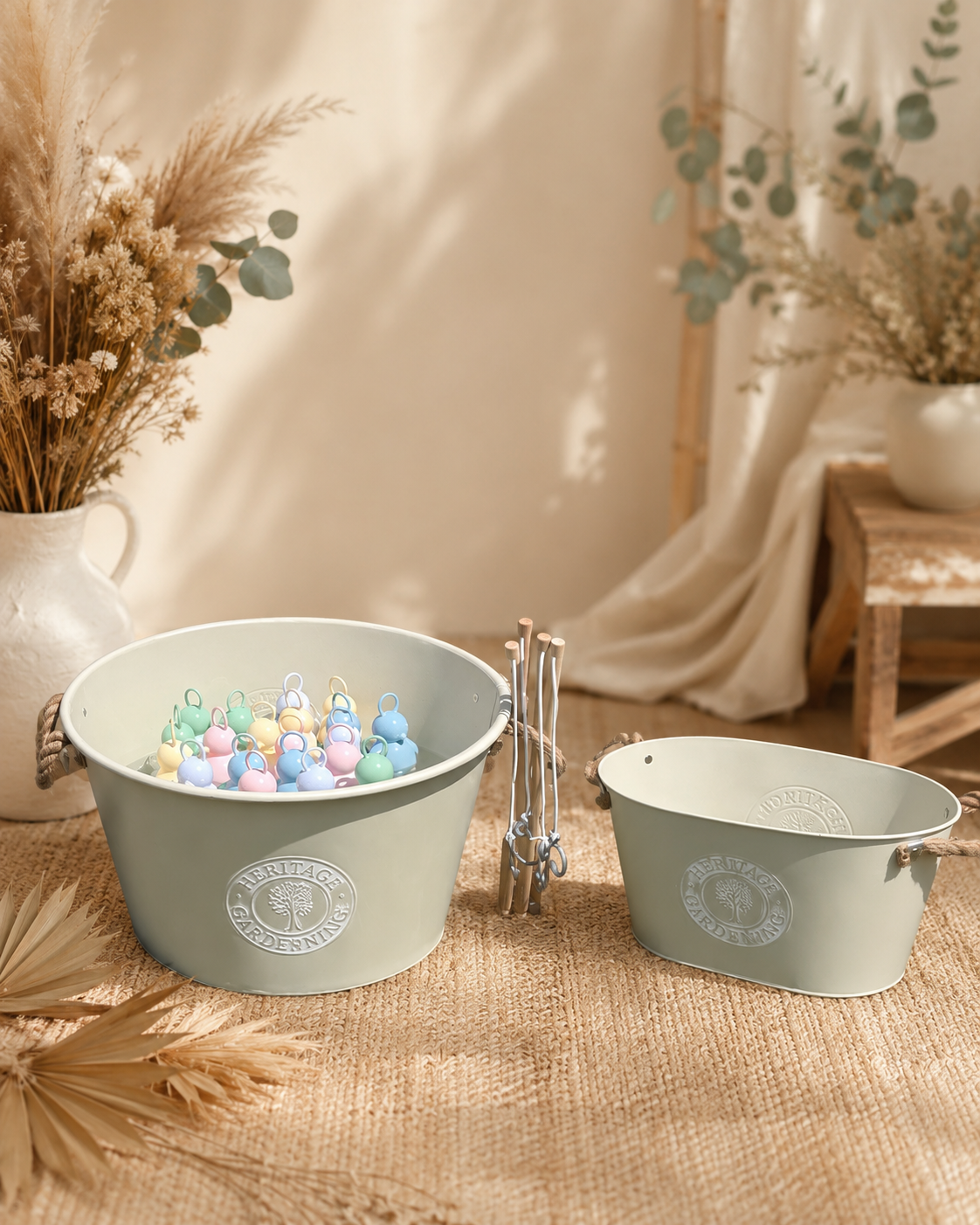 Deux seaux en plastique gris clair avec logo 'Heritage Gardening' remplis de petits objets colorés, entourés de décorations naturelles et d'une ambiance chaleureuse avec des fleurs séchées et un tissu léger en arrière-plan.