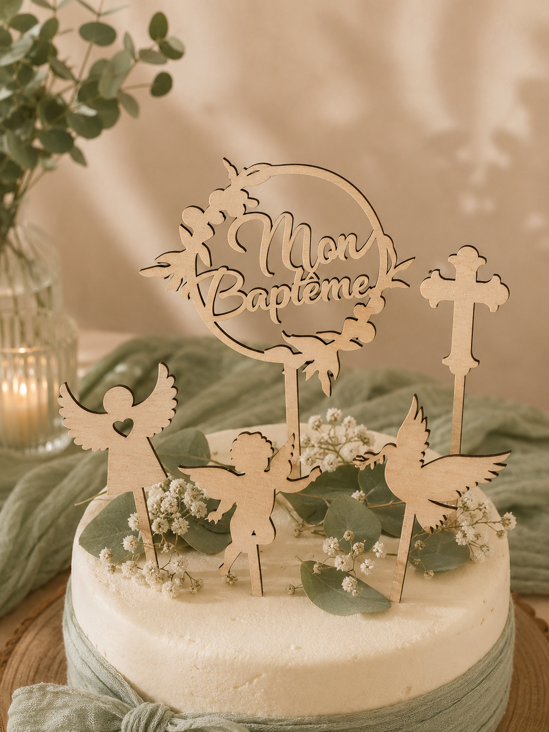 Gâteau pour la célébration du baptême, décoré avec des figurines en bois représentant des anges, une croix, des oiseaux et un cercle avec le texte 'Mon Baptême', entouré de petites fleurs et de feuilles vertes.
